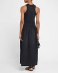 A person models the Racer Tank Dress by FRAME, standing with their back to the camera in a sleek, all-black ensemble. They hold a small clutch and wear sandals against a plain white background, perfectly capturing effortless style and simplicity.