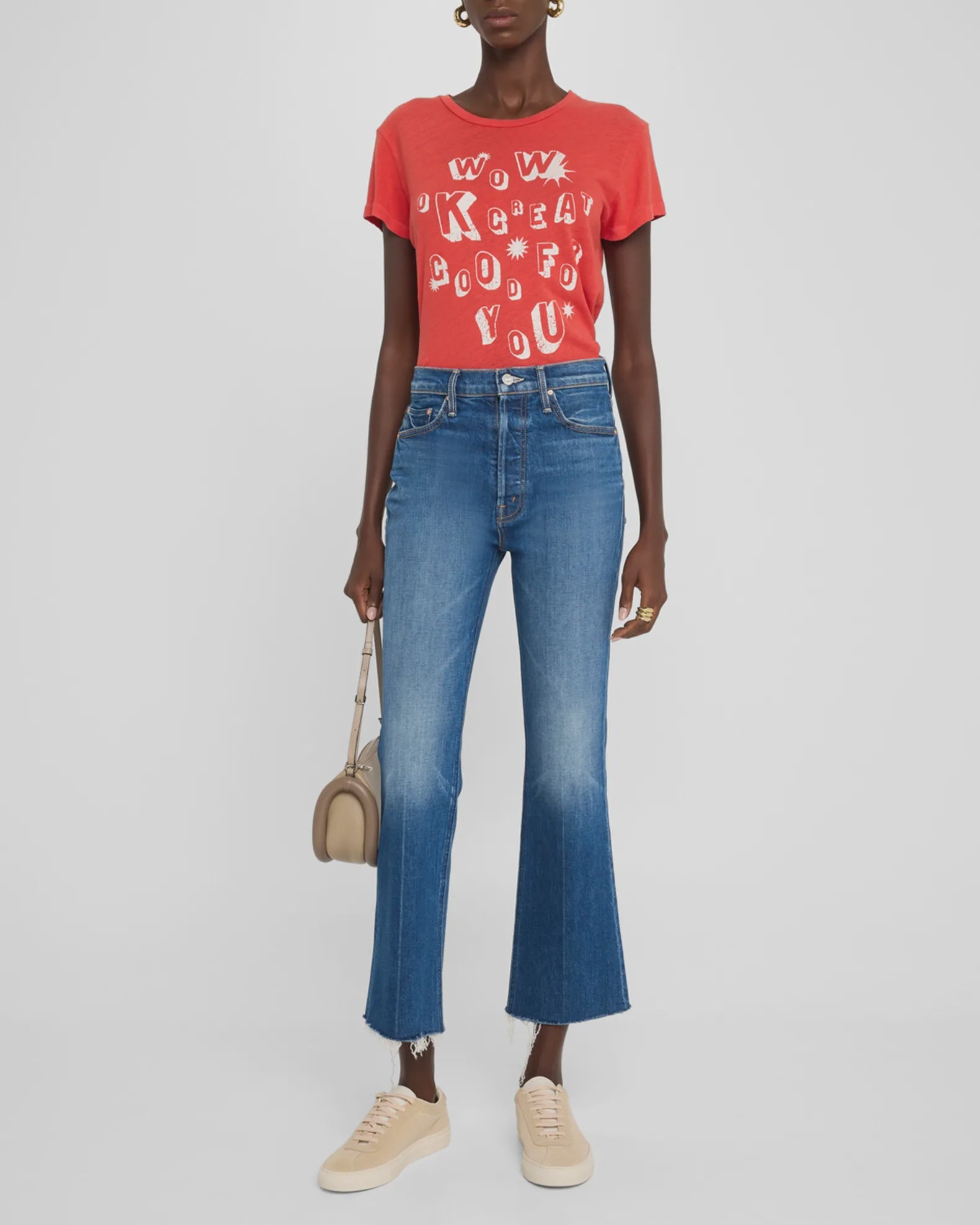 A person wearing a red T-shirt with white text and graphics, blue "The Tripper Ankle Fray" high-rise jeans by Mother featuring a frayed hem and five-pocket design, and beige sneakers stands holding a beige handbag. The background is plain white.