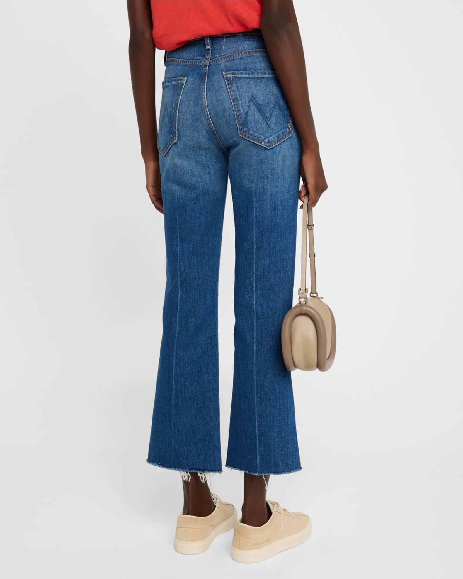 A person wearing a red shirt and The Tripper Ankle Fray high-rise blue flare jeans by Mother, designed with frayed hems and made from cotton/elastane, is holding a beige handbag. They are also sporting light-colored sneakers against a plain white background.