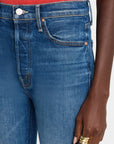 A person is dressed in The Tripper Ankle Fray jeans by Mother, characterized by their high-rise, ankle-length blue denim with distinctive orange stitching. The cotton and elastane blend ensures both comfort and a great fit. Complementing the look is a subtly visible red top, while multiple gold rings embellish the hand resting near the pocket.