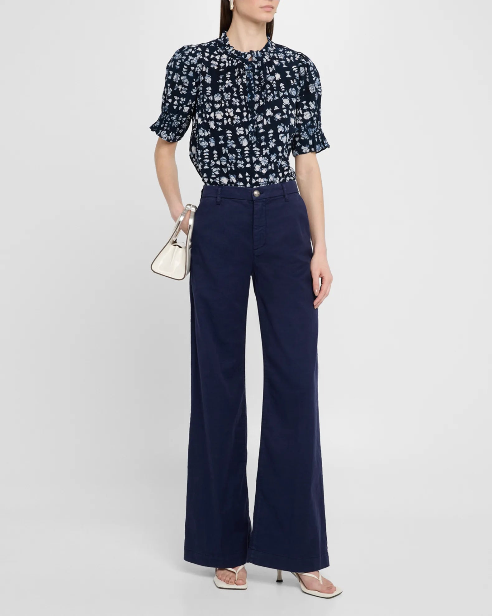 Woman wearing the Apiece Apart Los Altos Top in navy with an abstract print, tucked into high-waisted wide-leg navy pants, paired with a small white handbag and white heeled sandals.