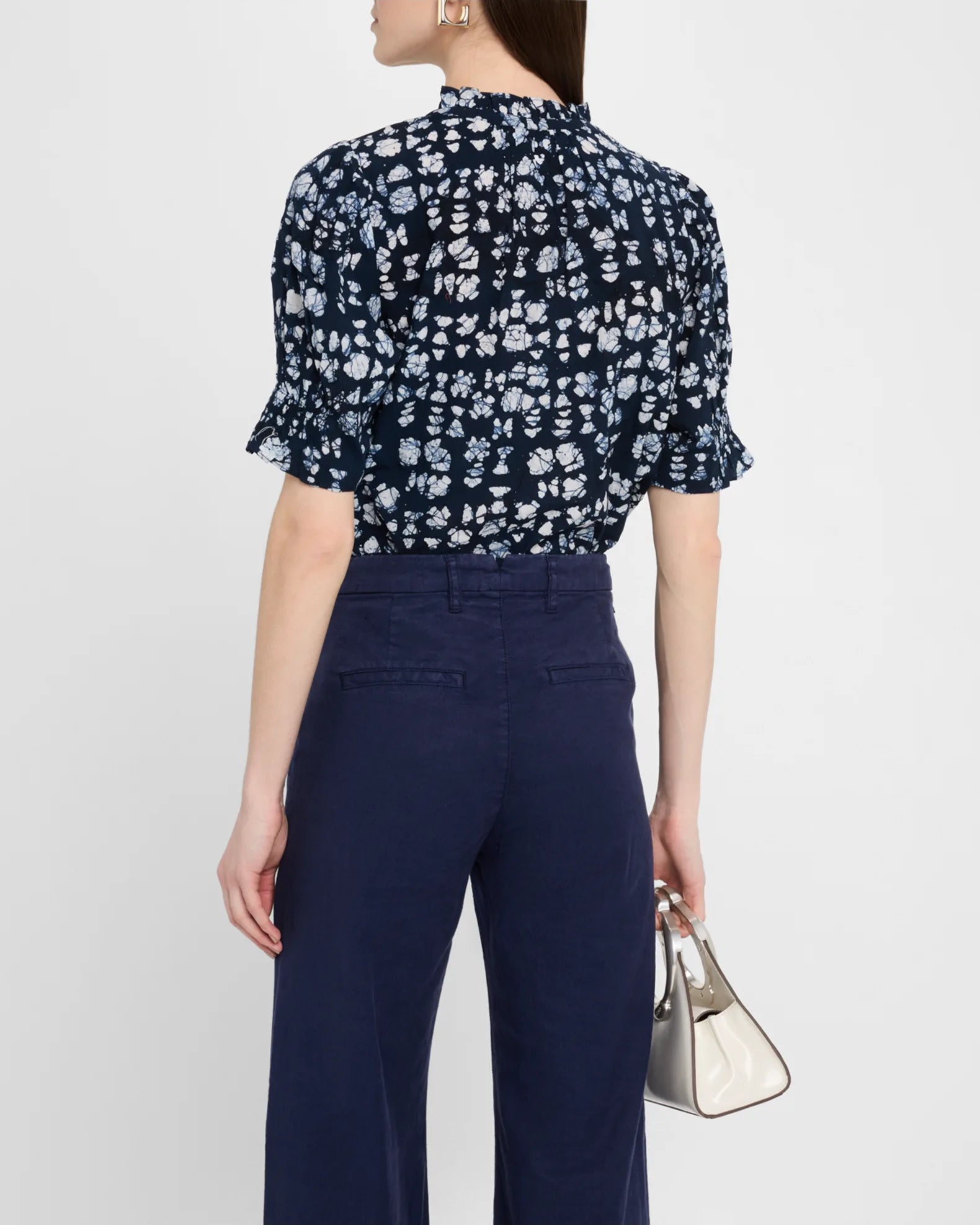 A woman, with straight brown hair down, faces away wearing Apiece Apart’s Los Altos Top— a navy floral puff-sleeve blouse tucked into high-waisted navy trousers. She holds a small white handbag and wears statement earrings.