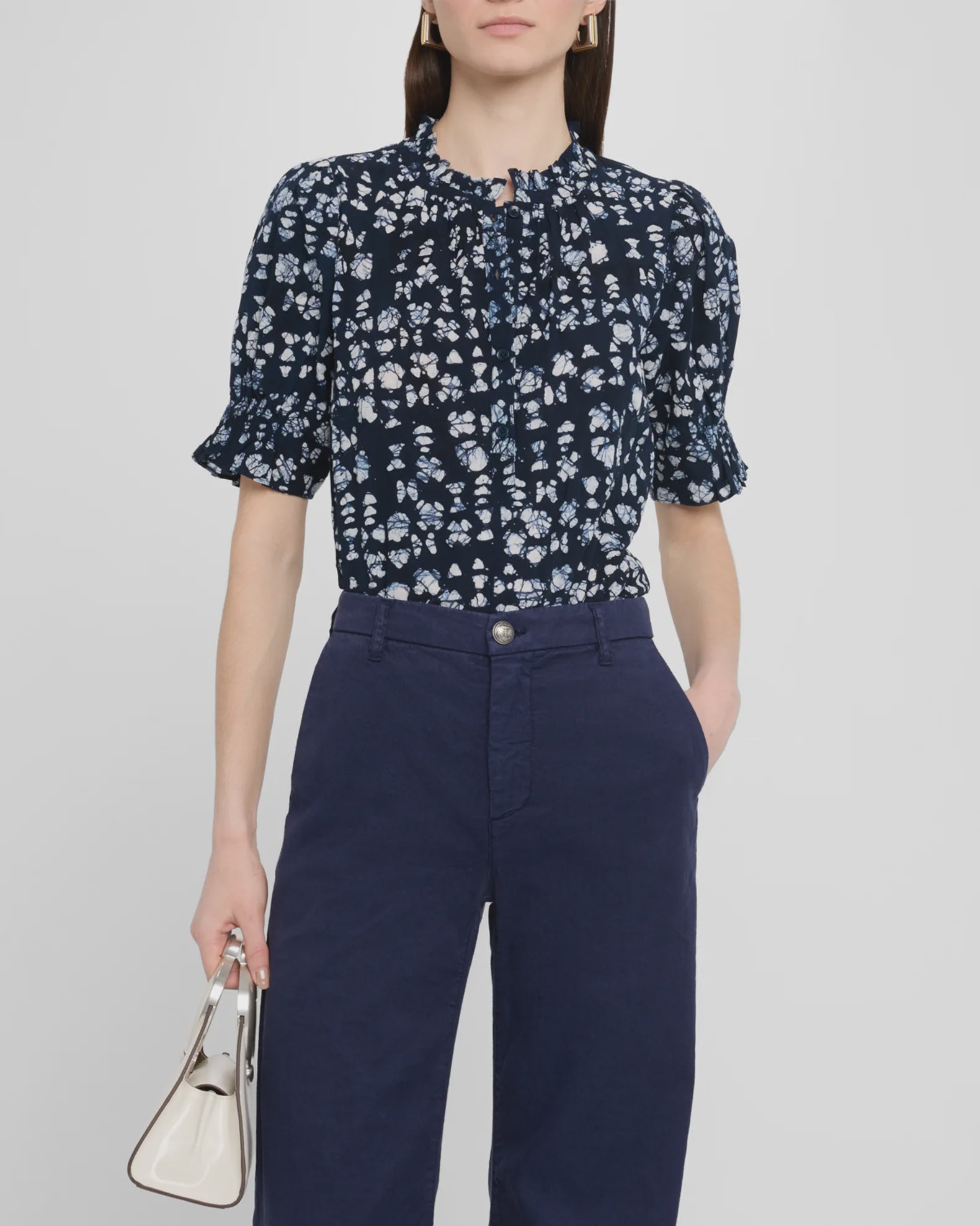 A woman stands against a plain light background wearing the Apiece Apart Los Altos Top, a navy blue short-sleeve blouse with a crew neckline and floral print, tucked into high-waisted navy pants and holding a small white handbag.