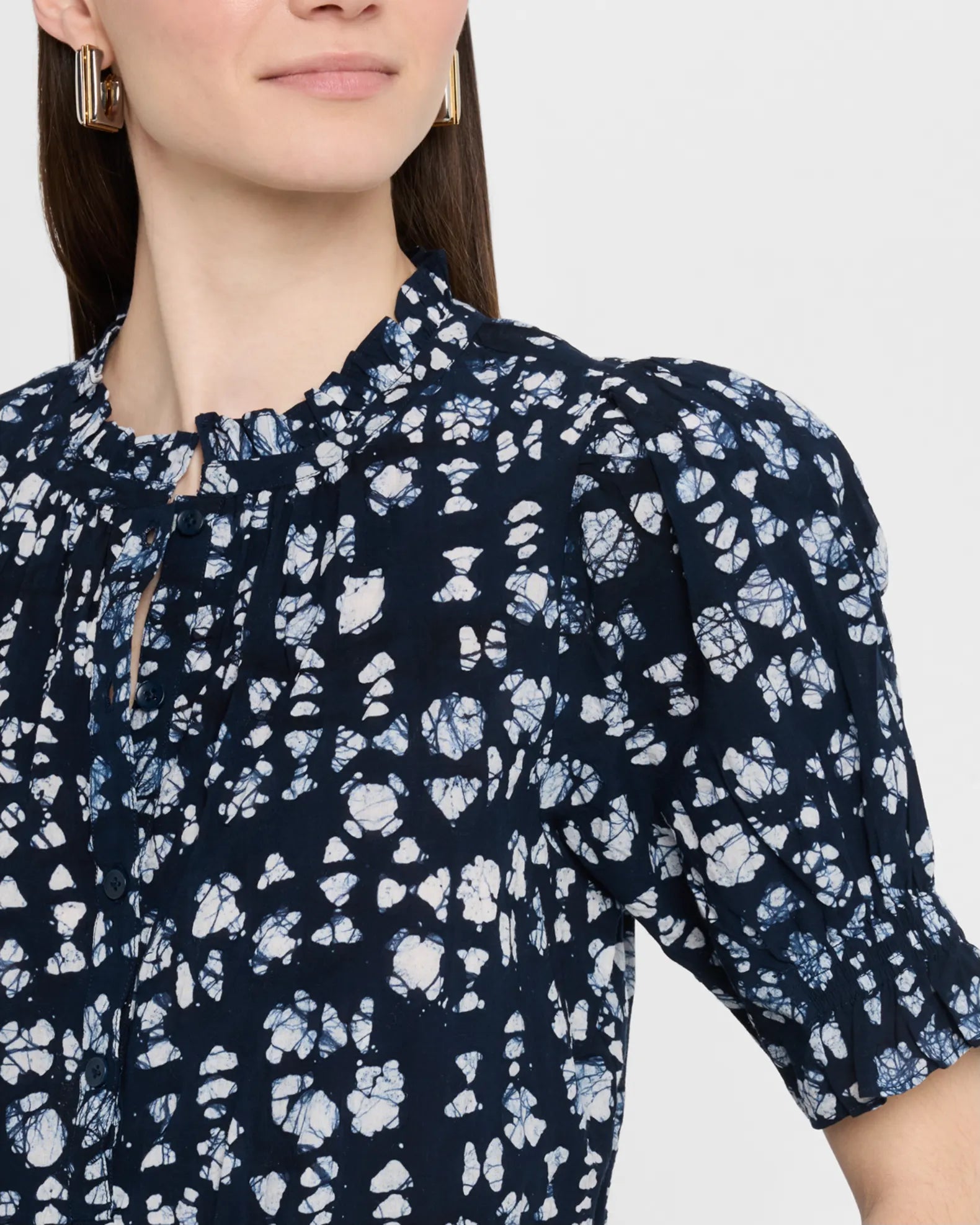 A woman wears the Apiece Apart Los Altos Top in navy blue with an abstract print, ruffle collar, and puffed short sleeves. She accessorizes with gold rectangular earrings. Only her head and upper torso are shown.