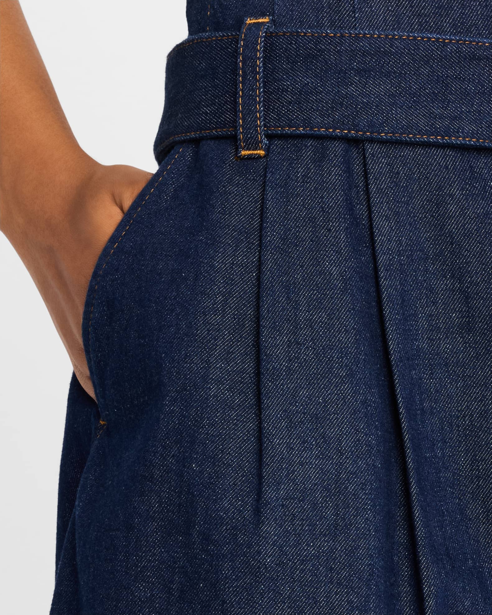 Close-up of a person wearing FRAME Paper Bag Shorts in high-rise dark blue denim, casually placing their hand inside the pocket. These shorts feature a pleated design with yellow stitching, offering style and comfort against a plain white background.