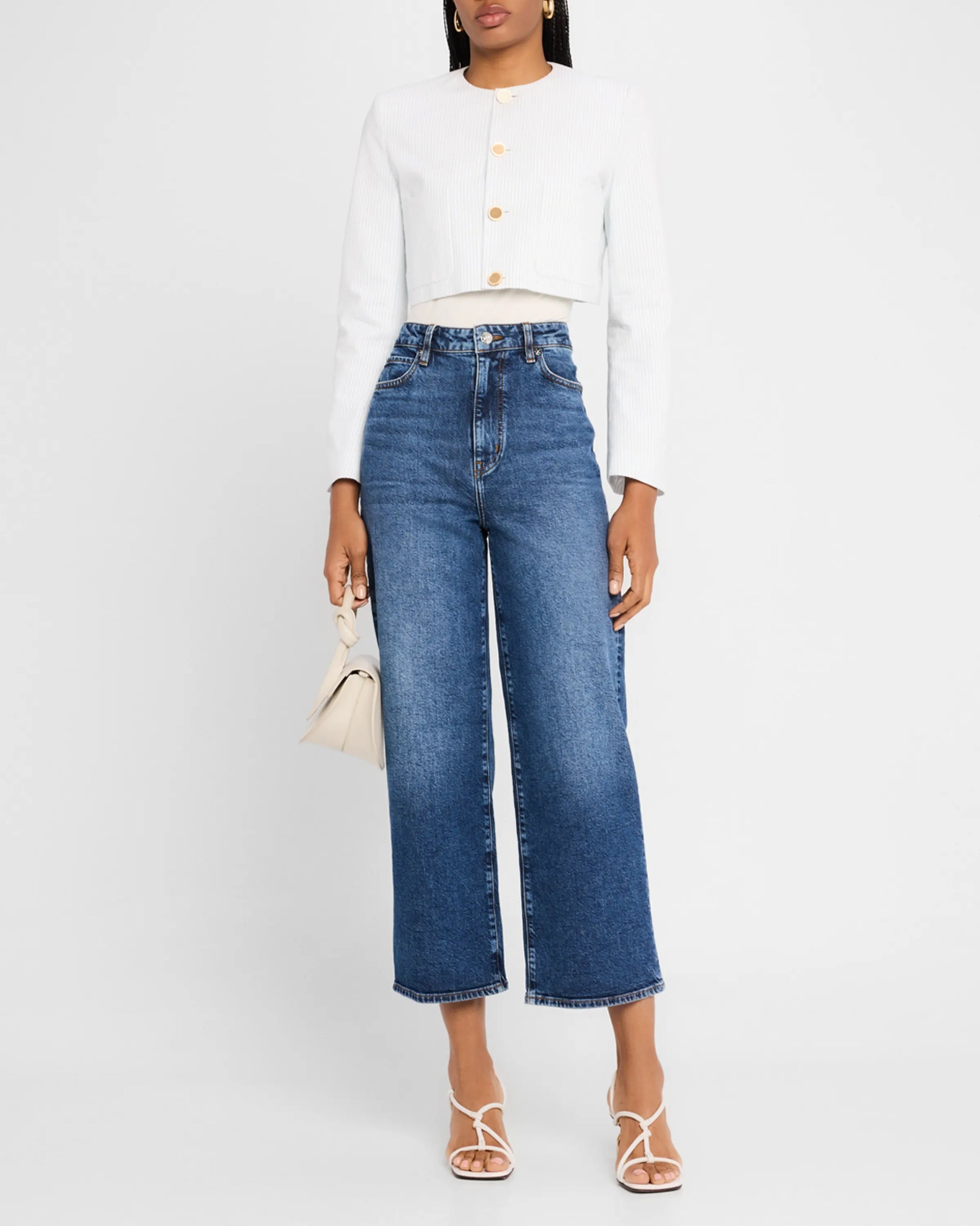 A woman wears FRAME's The Seersucker Jacket, styled with high-waisted wide-leg blue jeans, white strappy sandals, and a beige handbag.
