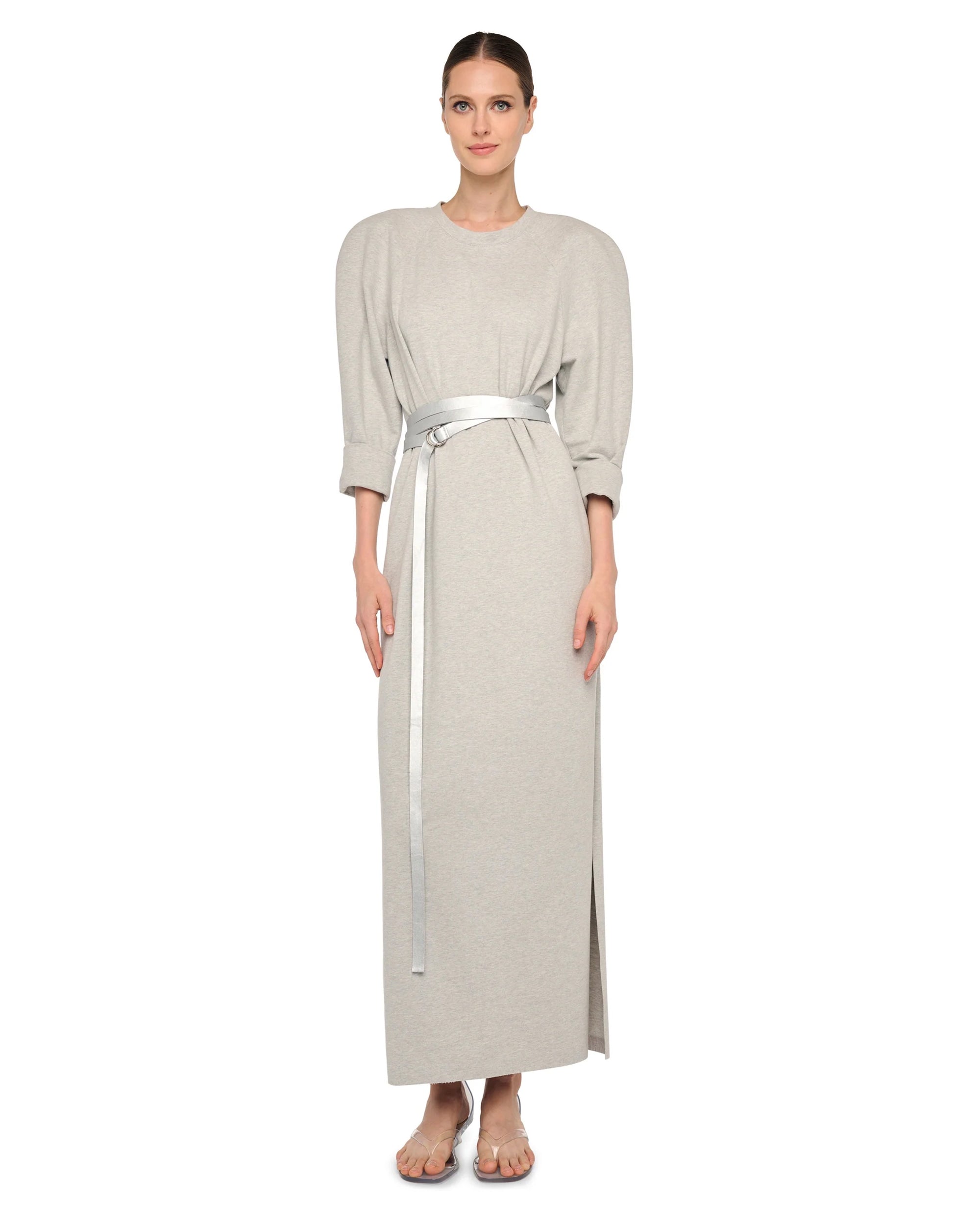 A woman stands barefoot against a white background wearing the Norma Kamali Bomber Long Dress—a vintage-inspired, light gray dress with three-quarter sleeves, subtle puffed shoulders, and a shiny silver belt at the waist.