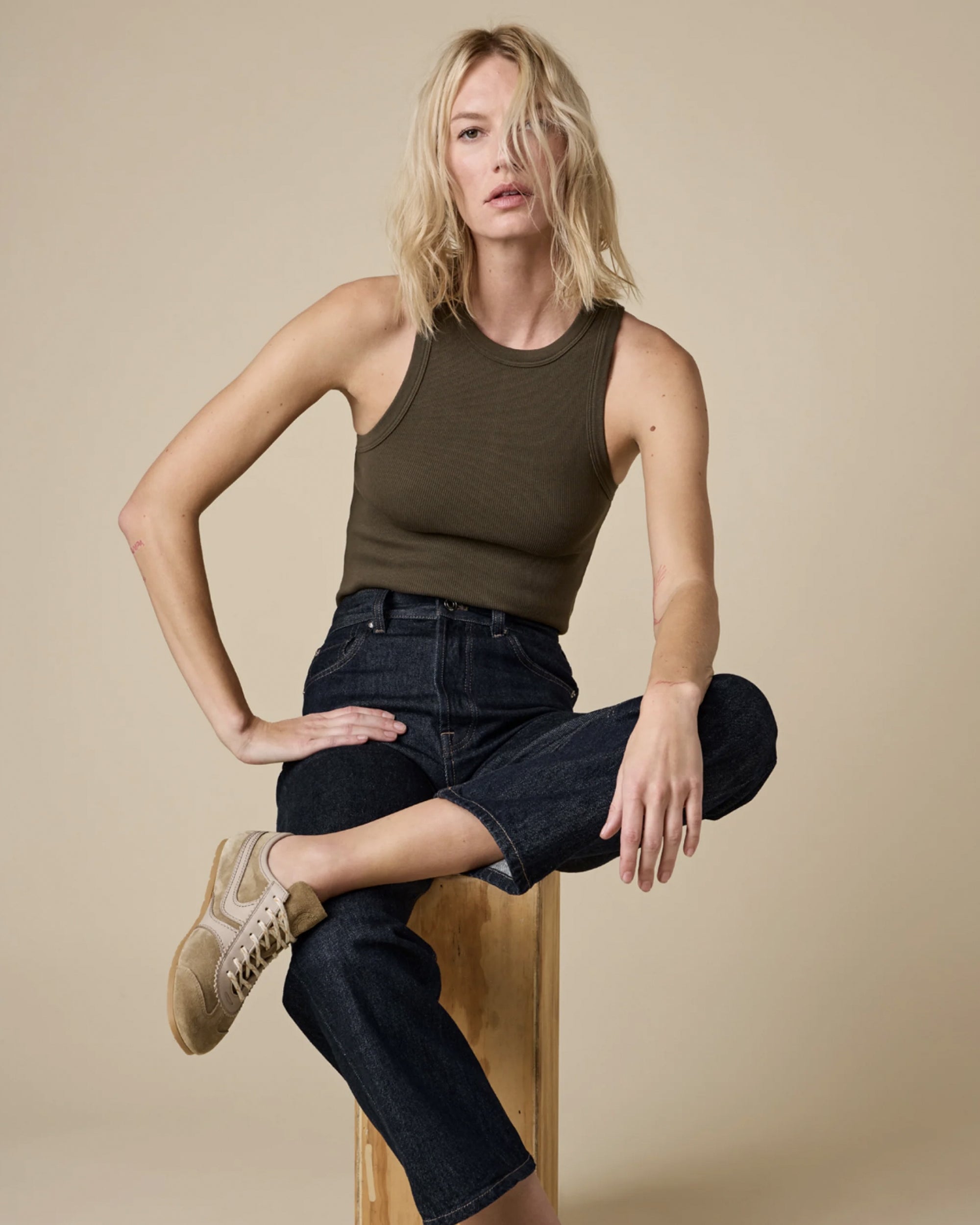 A woman with shoulder-length blonde hair wears Sold Out NYC's best-selling "The Not So Basic Tank" in dark olive, paired with dark high-waisted jeans and beige sneakers, as she sits on a wooden stool against a plain beige backdrop.