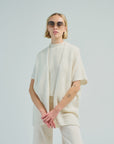 A person with short blonde hair, round sunglasses, a textured white top, the LaTierra Nube Vest, a loose cream cardigan, and light pants stands against a plain light background with arms loosely crossed.