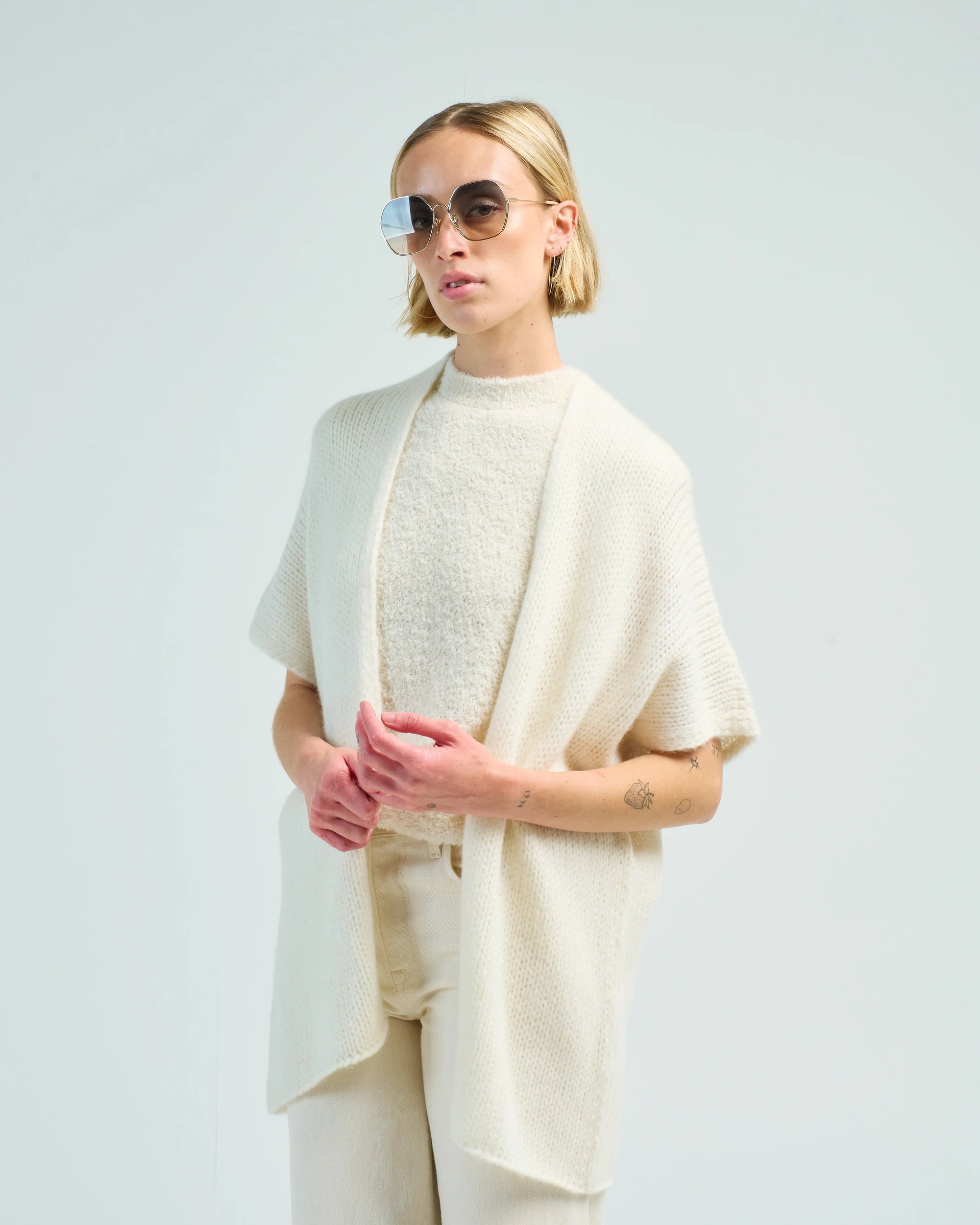A woman with short blonde hair and large sunglasses wears the LaTierra Nube Vest over a textured white top and cream pants, standing confidently against a plain light background.