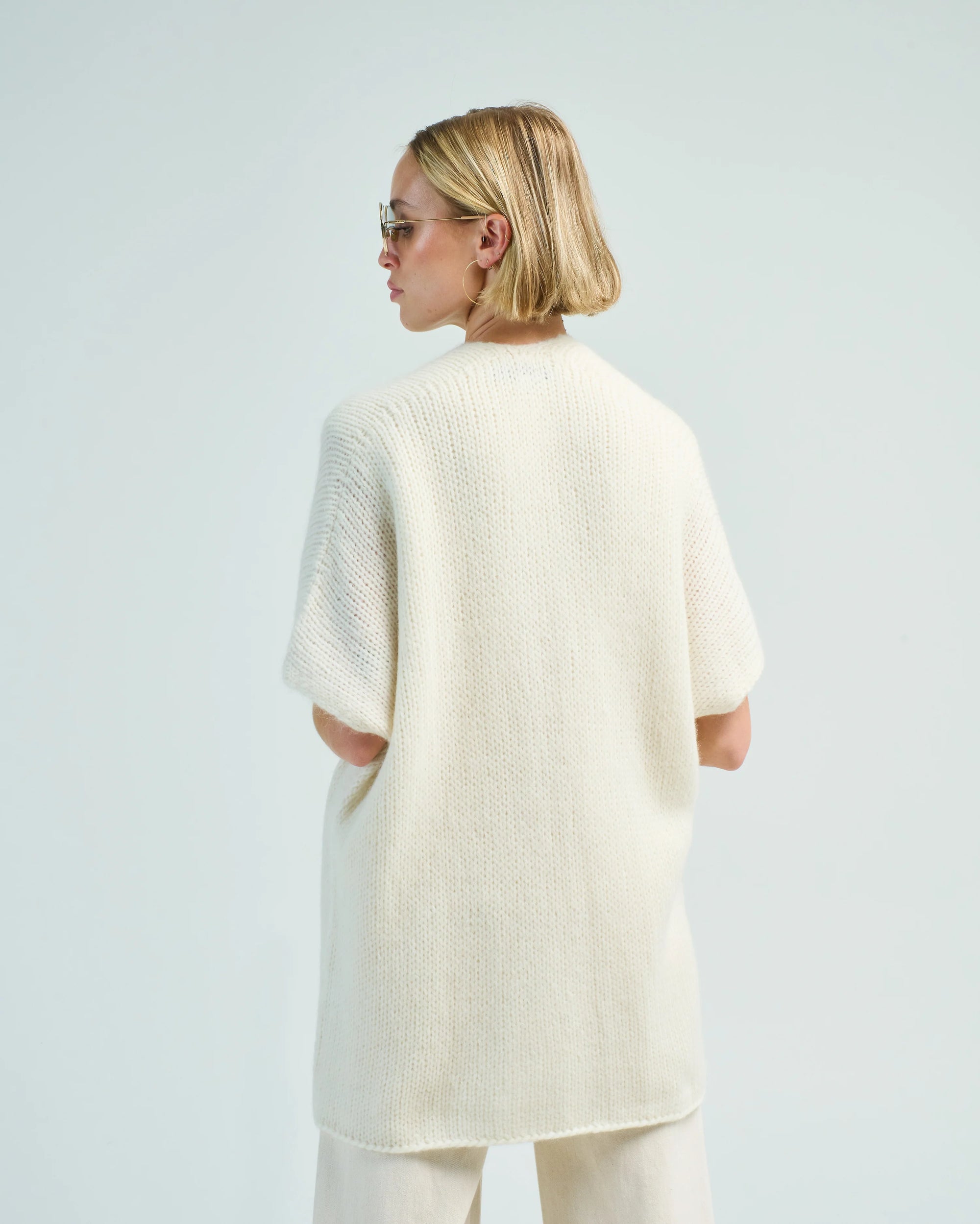 A person with short blonde hair and sunglasses wears the LaTierra Nube Vest layered over an off-white oversized sweater and cream pants, standing with their back to the camera against a plain light background.