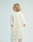 A person with short blonde hair and sunglasses wears the LaTierra Nube Vest layered over an off-white oversized sweater and cream pants, standing with their back to the camera against a plain light background.