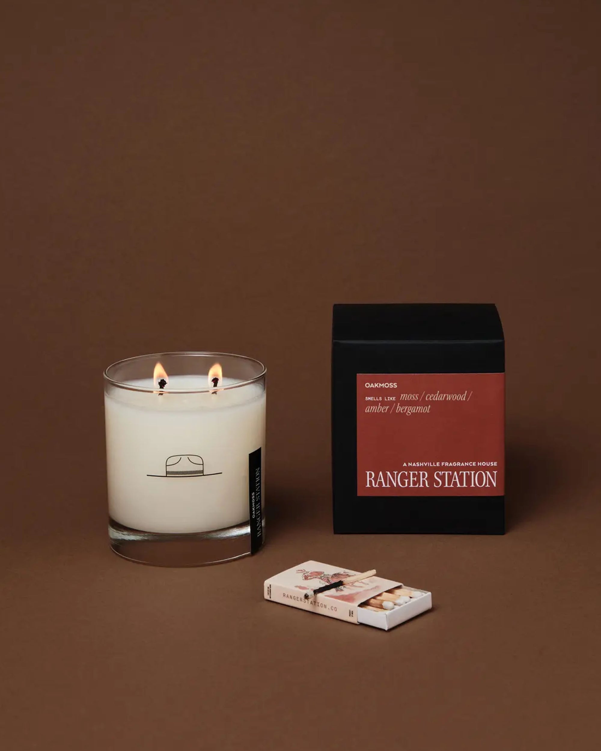 A lit Ranger Station Candle with double wicks in a glass jar featuring a hat logo sits beside its black and red box and a matchbox on a brown surface, filling the air with subtle cedarwood and oakmoss notes.