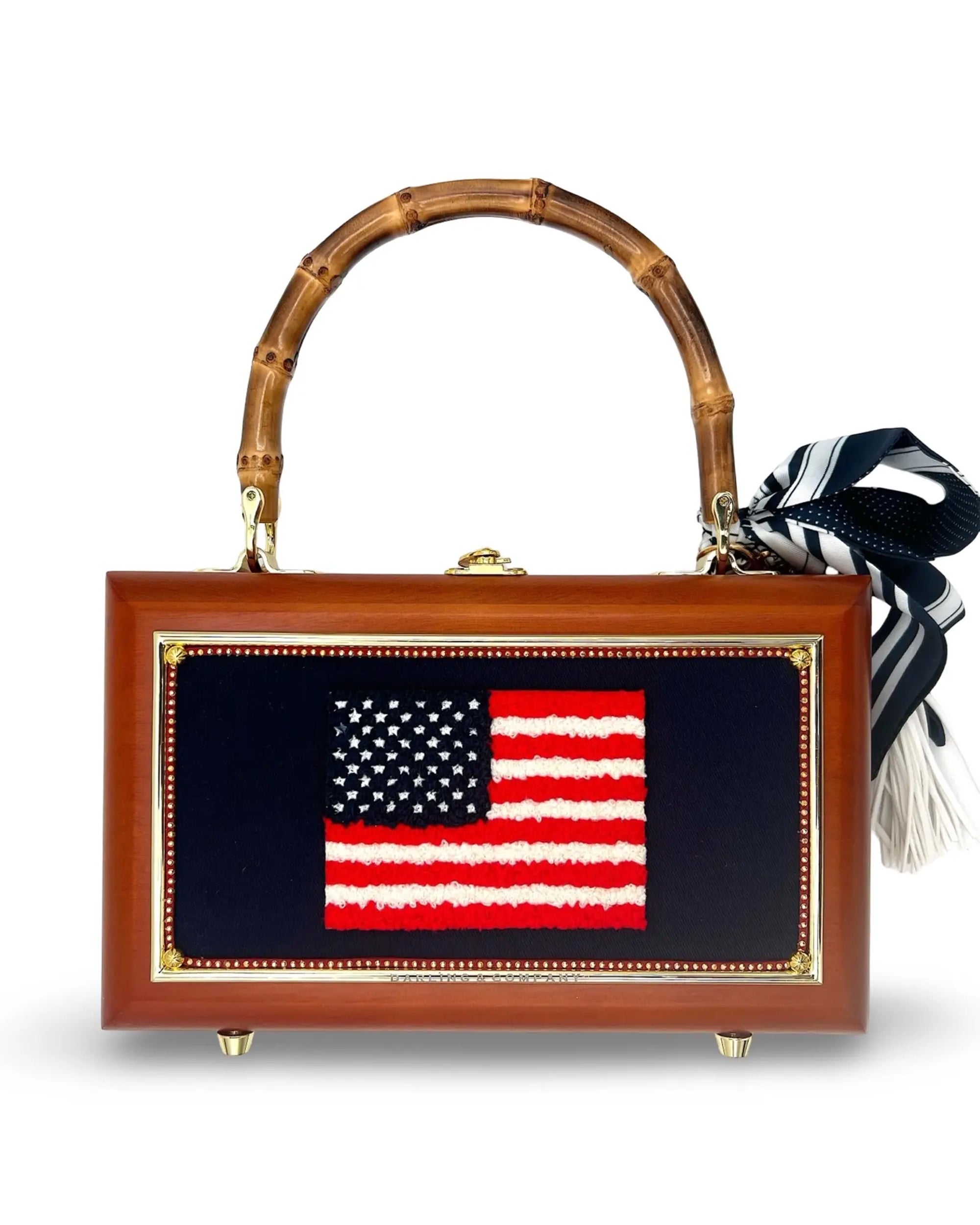 The Grand Old Flag Bag by Faire is a hand-finished designer wooden box handbag featuring a textured chenille American flag patch, bamboo handle, and a black-and-white striped scarf tied to the handle.