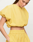 A woman is wearing a yellow, short-sleeved Opihi Bubble Sleeve Crop Top by Mikoh and matching high-waisted pants with an elastic waistband. The top has a loose, puffy design, made of 100% cotton, and the pants have side pockets. She has light brown hair tied back and is looking down. The background is plain white.