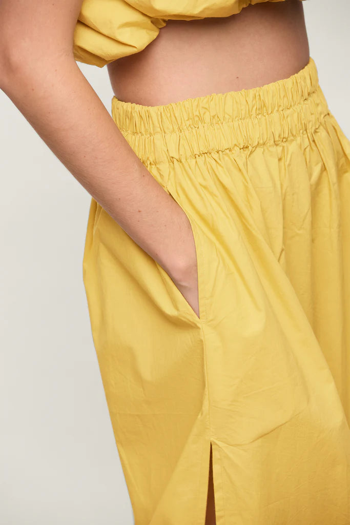 Close-up of a person wearing the Mikoh Delia Maxi Skirt with an elastic waistband. Their right hand is in the skirt pocket. The skirt features a high slit, and the fabric appears to be lightweight. The person is standing against a plain, light-colored background.