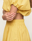 A person is shown from the shoulders down, wearing a yellow two-piece outfit featuring the Mikoh Opihi Bubble Sleeve Crop Top with an elastic waistband. The person's arms are crossed, with one hand gently holding the other arm. The background is plain and light-colored.