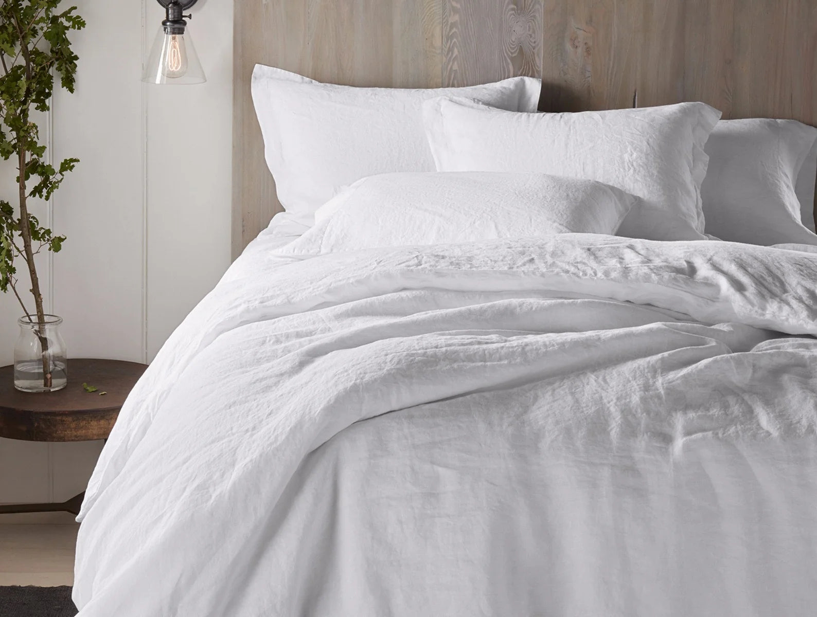 A cozy bed with fluffy white linens and pillows features the Coyuchi Inc Org Relaxed Linen Duvet in Alpine White, crafted from organic linen and low-impact dyes, set against a light wooden headboard beside a dark round bedside table.