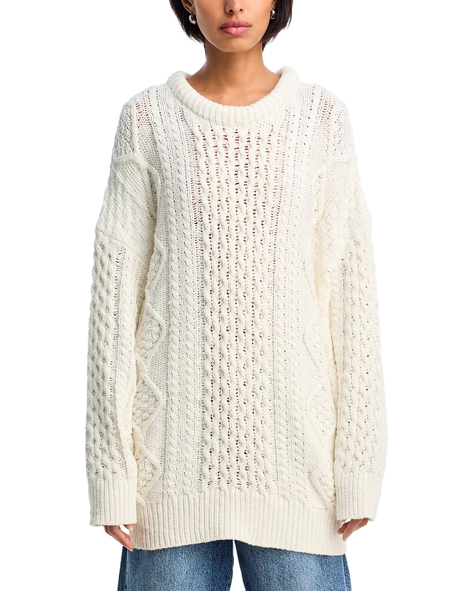 A person wears the Denimist OVERSIZED ARAN SWEATER—a loose, chunky-knit cream cotton piece with cable patterns and ribbed details—paired with blue jeans. The cropped image emphasizes the sweater’s cozy, oversized fit.