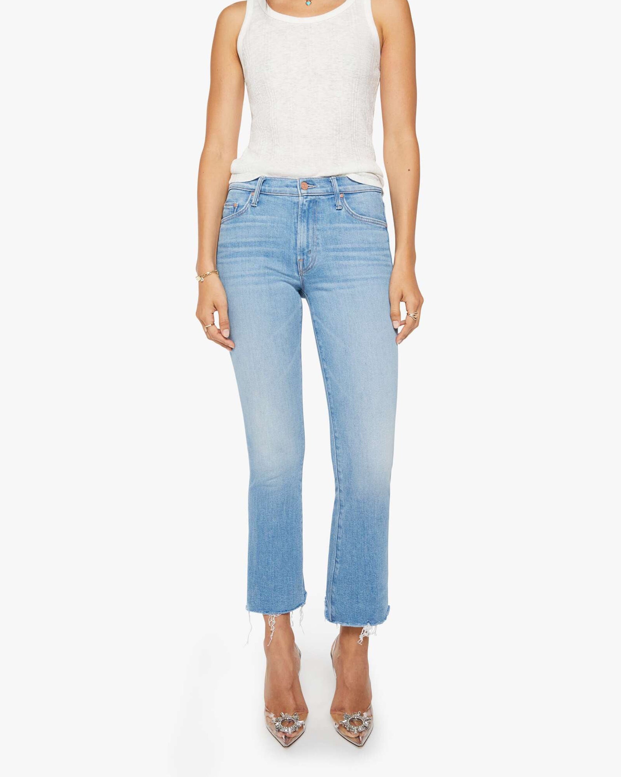 A person wears Mother’s The Outsider Flood Fray light blue straight-leg jeans with frayed hems, a sleeveless white top, and pointed-toe clear heels, standing against a white background.
