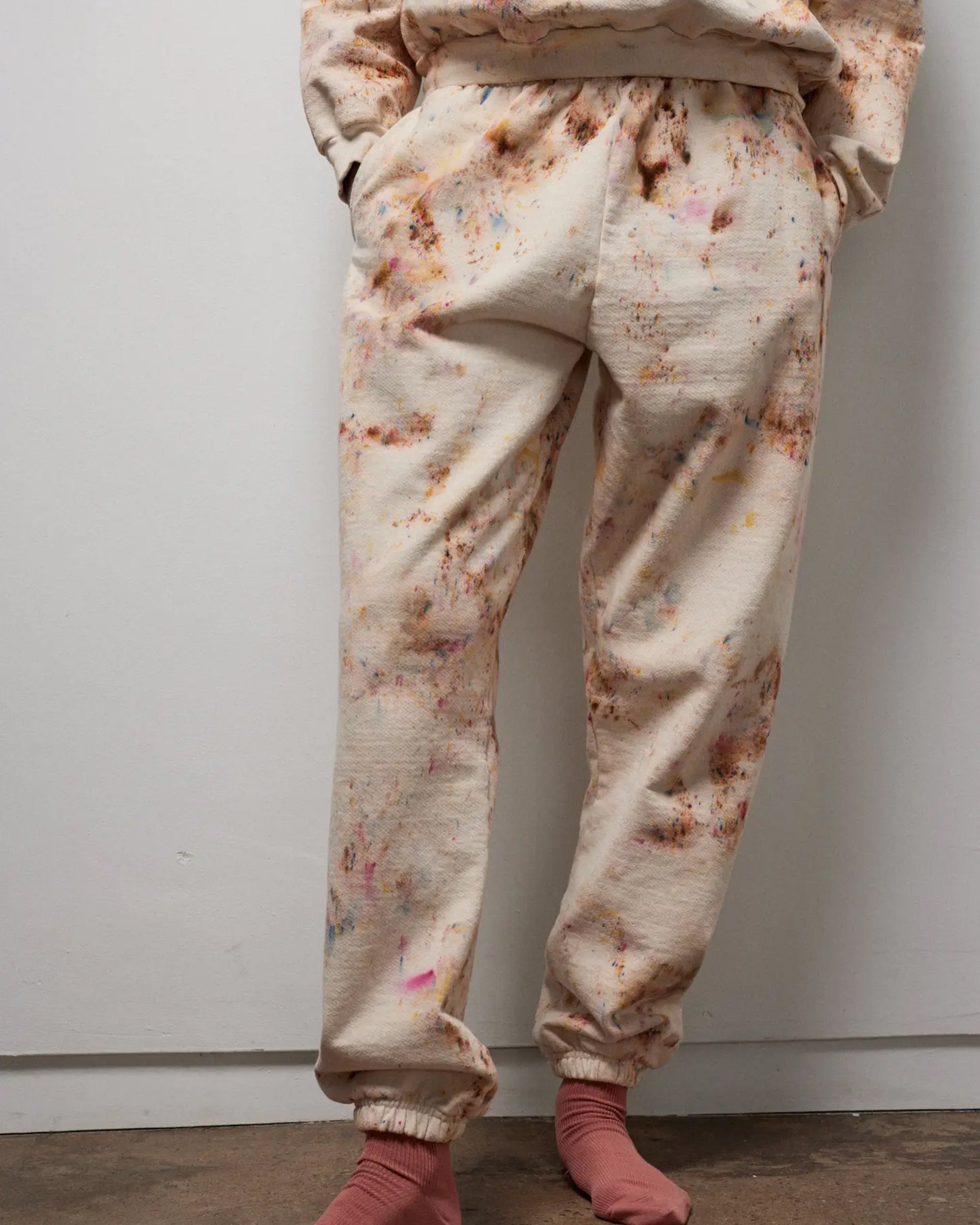A person models the Faire Paint Splatter Sweats—cream-colored heavyweight fleece loungewear with a colorful paint splatter design—indoors against a white wall, hands in pockets, paired with pink socks.