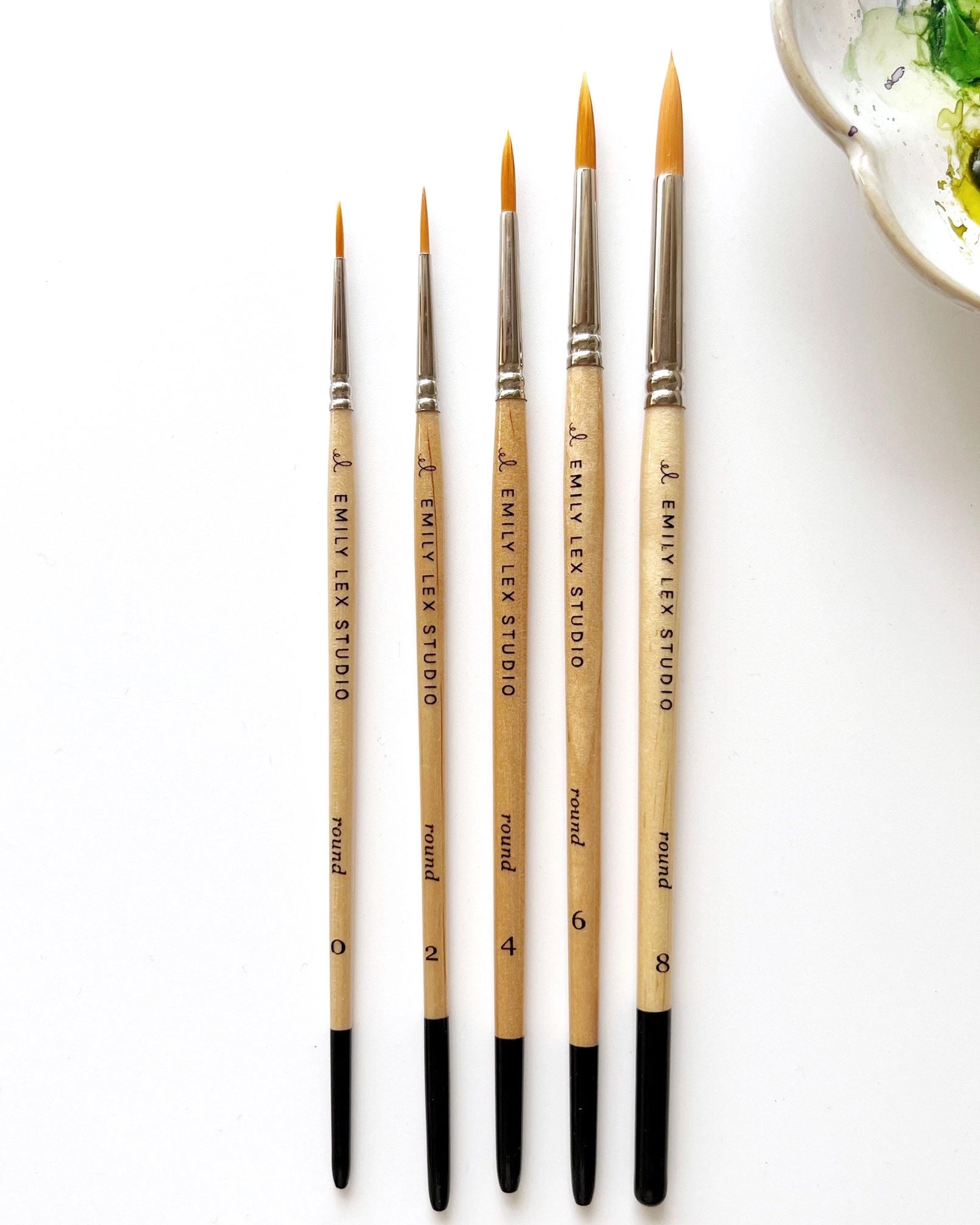 Five Faire Paintbrush Set round brushes, sizes 0, 2, 4, 6, and 8 with synthetic bristles and lacquered wood handles, are lined up on a white surface. A watercolor ceramic palette is partly visible in the top right corner.