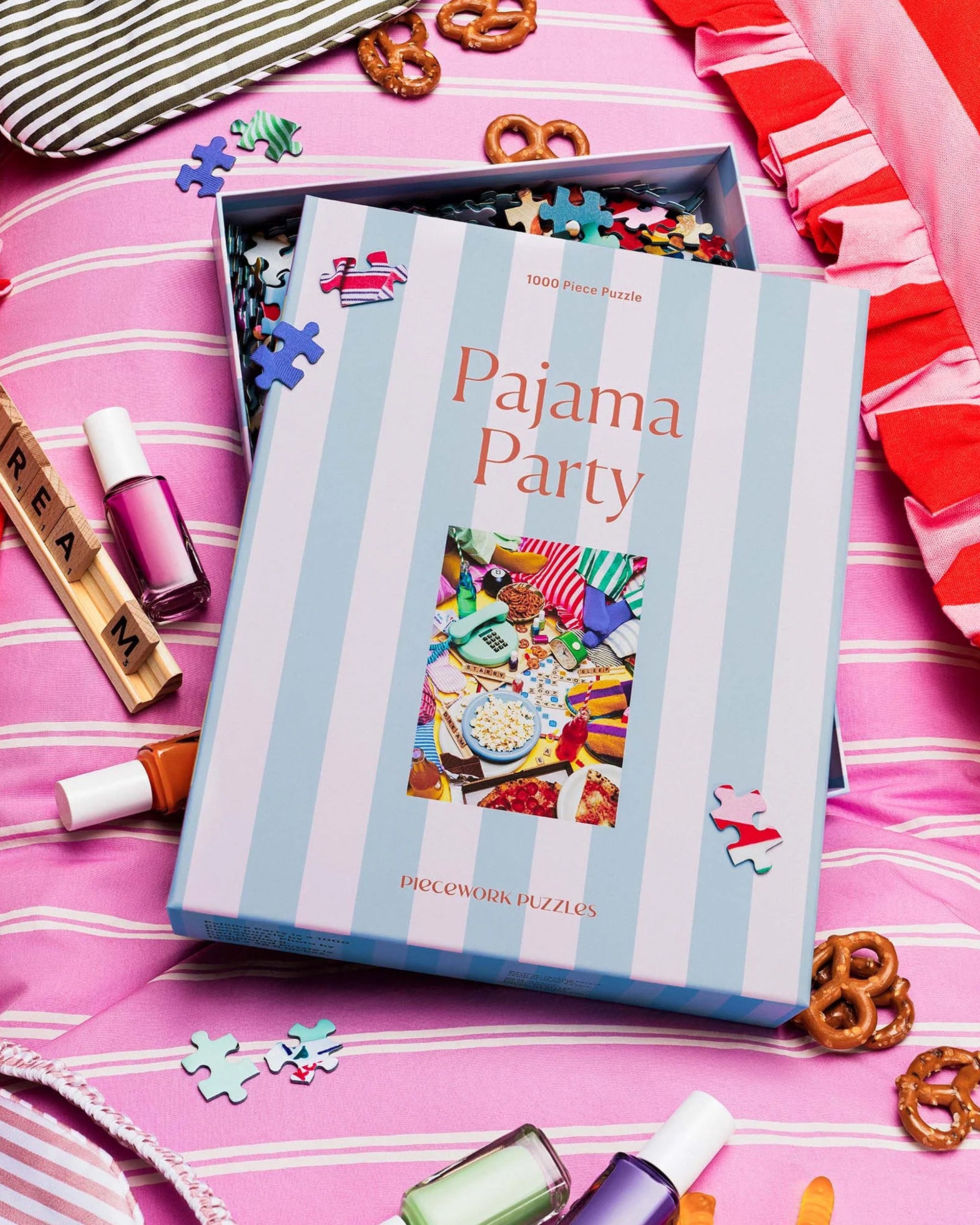 An open box of Faire’s 1000-piece Pajama Party Puzzle rests on a pink striped blanket, loose recycled-paper pieces scattered among nail polish bottles, pretzels, and a Scrabble rack spelling "DREAM.