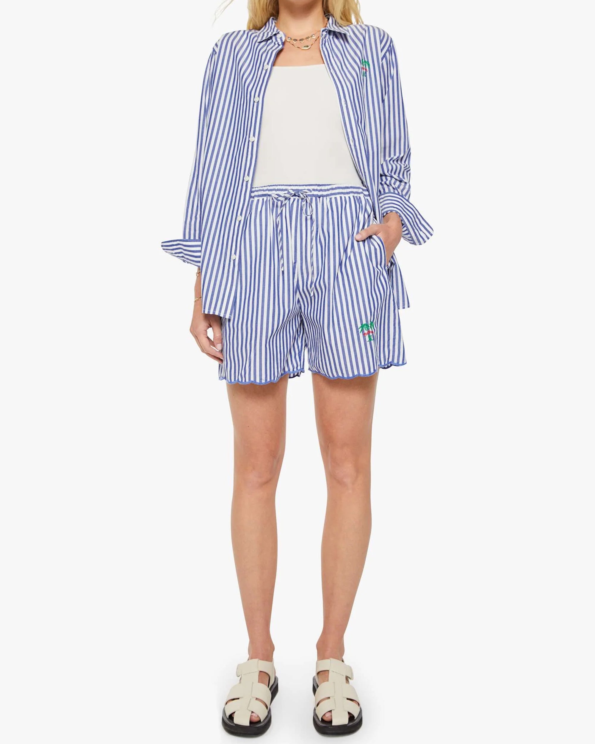A person wears the Mother Drawn FreeBaller Short—blue and white striped, lightweight cotton—with a matching button-up shirt over a white top. The shirt sleeves are rolled up, one hand is in a pocket, tan sandals finish the look. Head not shown.