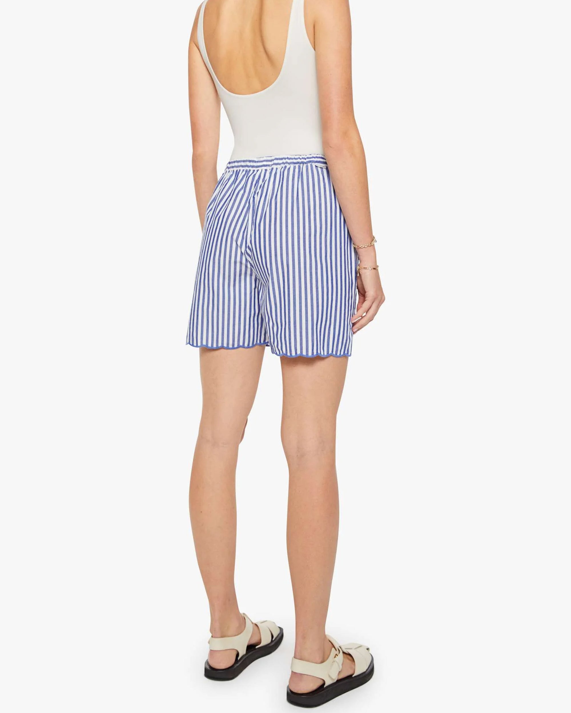 A woman faces away, wearing Mother’s The Drawn FreeBaller Short—blue and white striped with a scalloped hem—a sleeveless white top, white sandals with black soles, and a bracelet on her left wrist.