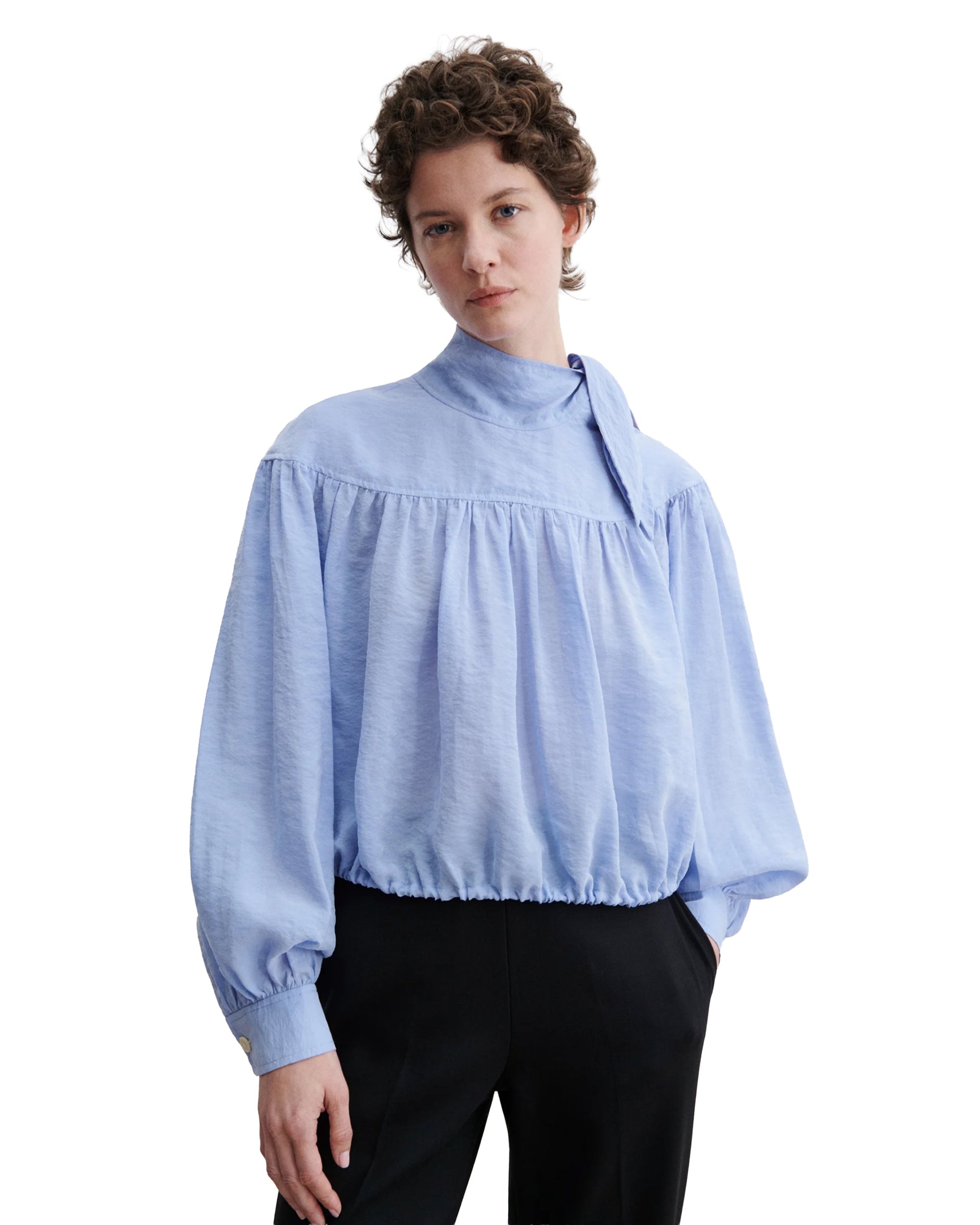 A person wears the Rachel Comey Parachute Top—a wrinkle-resistant, light blue, long-sleeve blouse with a tie collar and loose fit—paired with black pants, hands in pockets against a white background; ideal for travel.