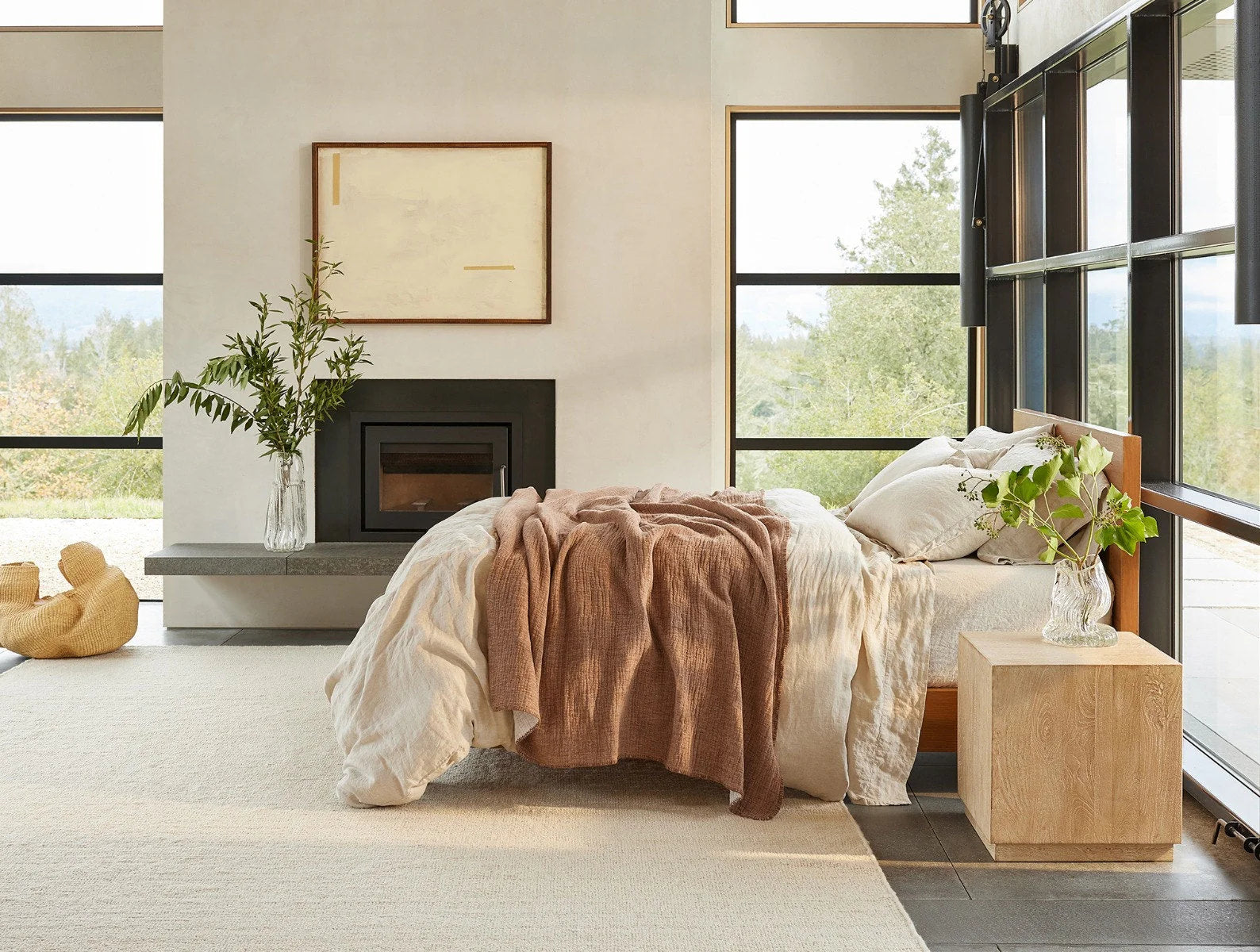 A cozy bedroom features a neatly made bed topped with the Cozy Cotton Org. Blanket by Coyuchi Inc, a wooden nightstand with greenery, a fireplace, large sunlit windows, and minimalist modern decor.