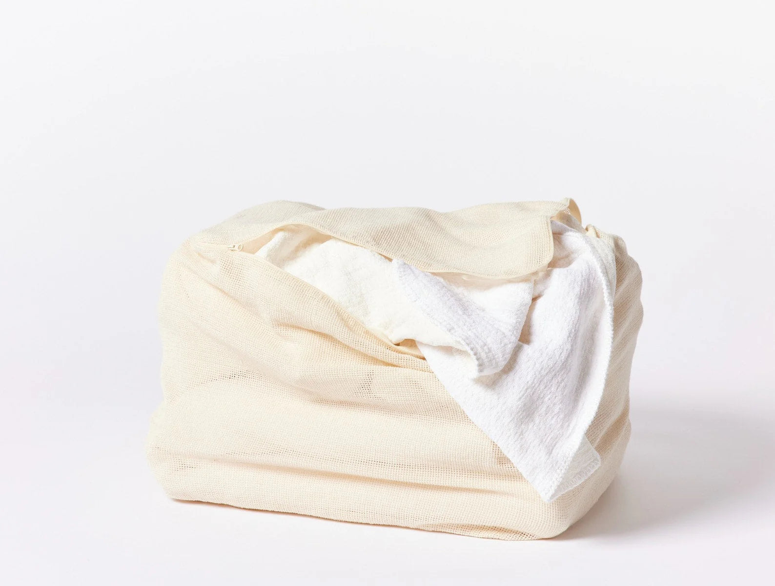 The Coyuchi Inc Organic Laundry Bag XL in beige, made from organic cotton mesh, sits against a white background, filled with white towels. One towel is partially draped over the edge of this natural laundry solution.