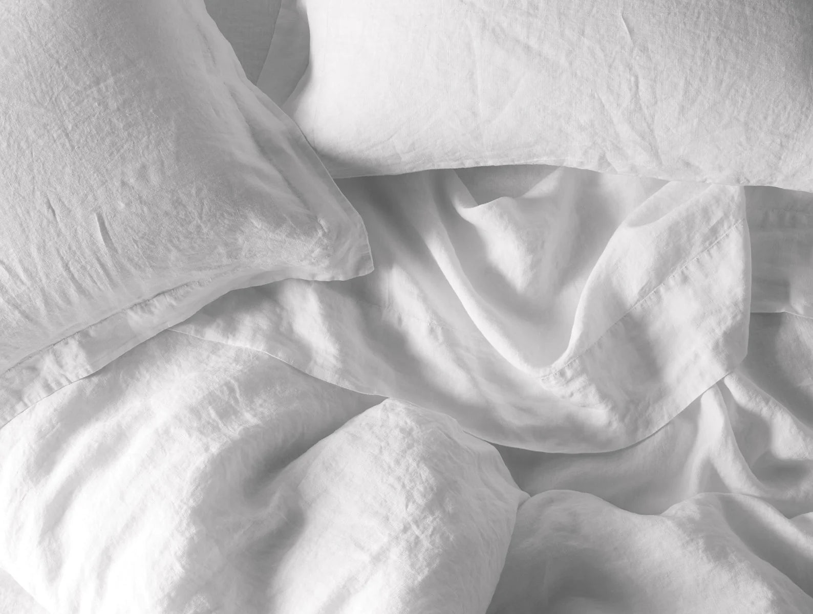 A close-up of Coyuchi Inc’s Org Relaxed Linen Duvet in Alpine White (Queen) with white organic linen sheets and pillows, softly rumpled and unmade, showcasing breathable fabric and low-impact dyed finish.