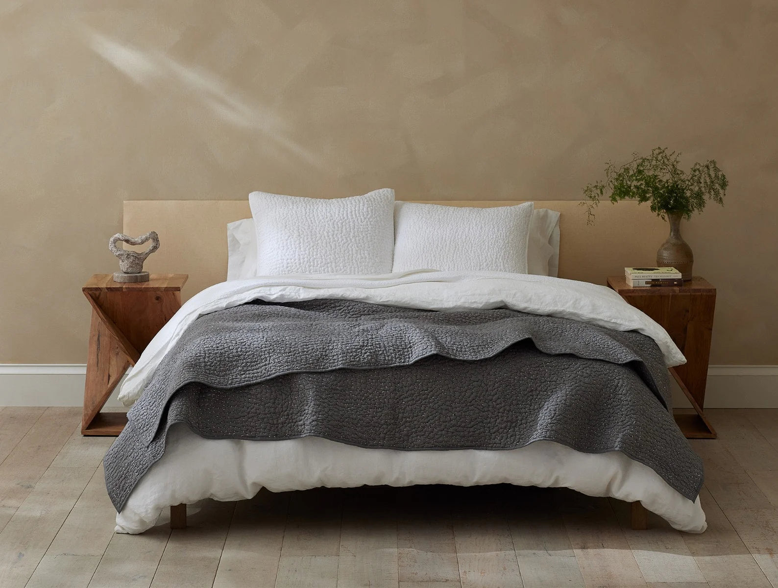 A neatly made bed with white pillows, a white sheet, and the Coyuchi Inc Pebbled Org Quilt in Queen Slate adds texture. Wooden nightstands hold a sculpture, potted plant, and books. Light wood floors and beige walls complete the serene room.