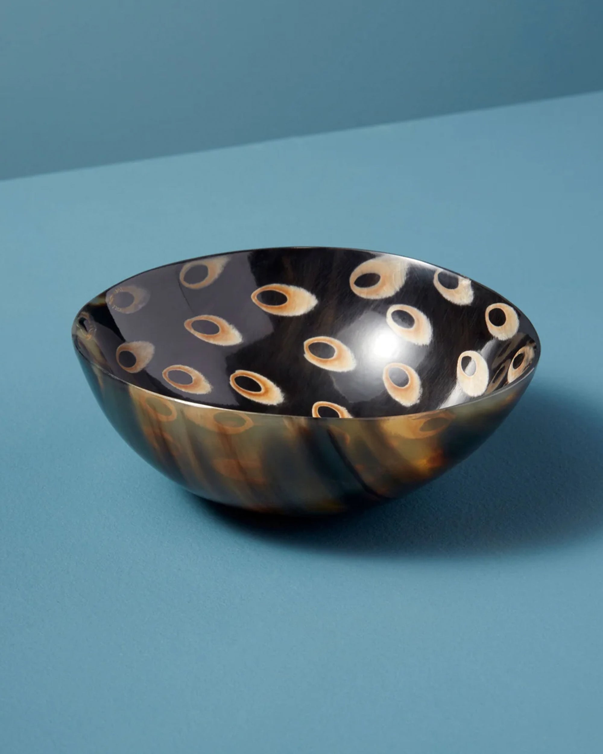 The Peacock Horn Bowl L by Be Home features a glossy, handcrafted finish with a dark marbled exterior adorned with tan and cream circular spots, shown on a light blue surface against a blue background.