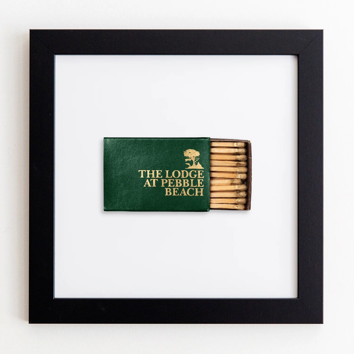 A framed acrylic display featuring a dark green book titled &quot;The Lodge at Pebble Beach&quot; with its spine exposed, revealing a series of matches integrated into the book design, against a white background by Match South&#39;s Art Square Black Frame.