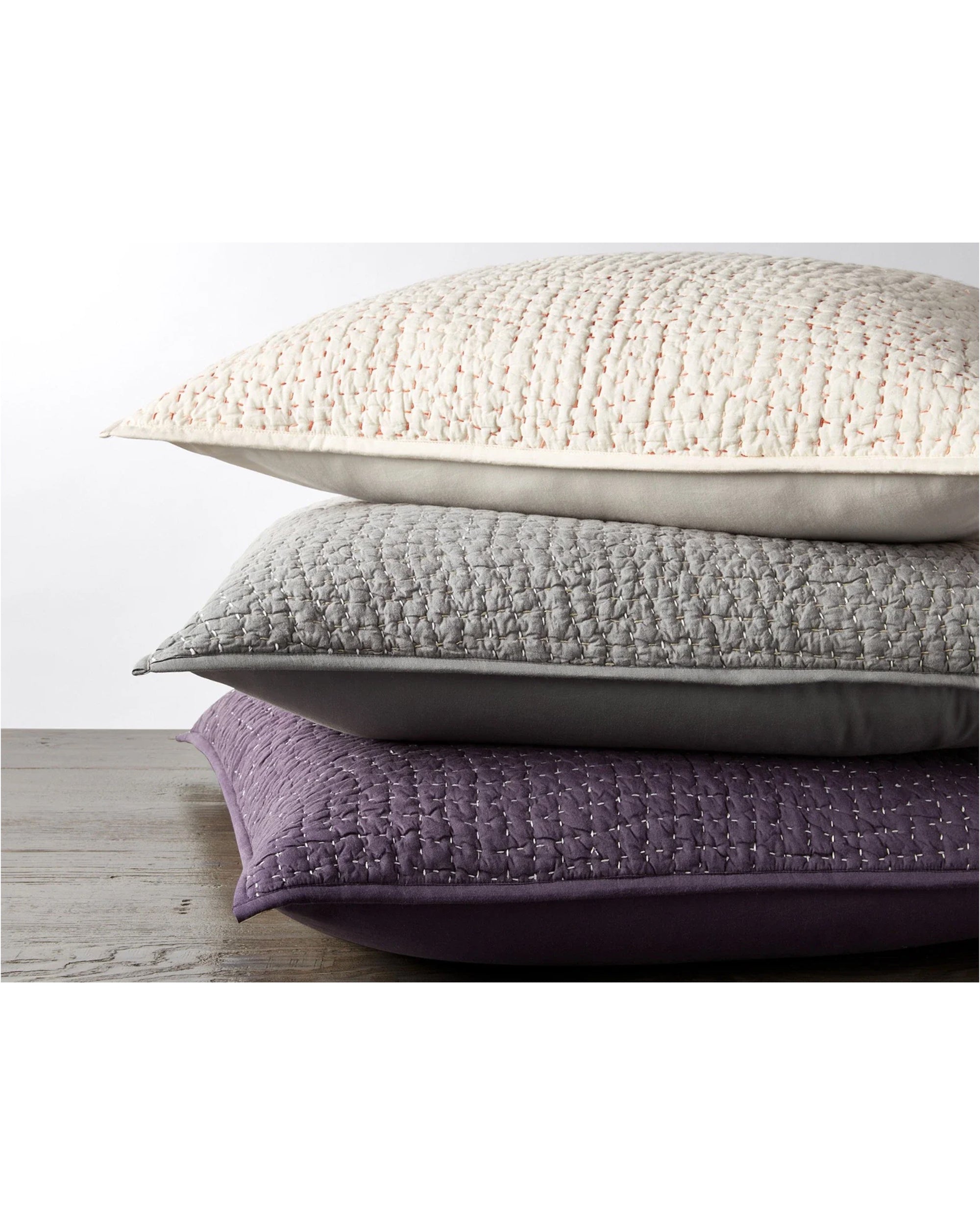 The Coyuchi Inc Pebbled Org Sham Euro Slate, crafted from soft organic cotton with a quilted texture, is shown stacked on a wooden surface against a white background.