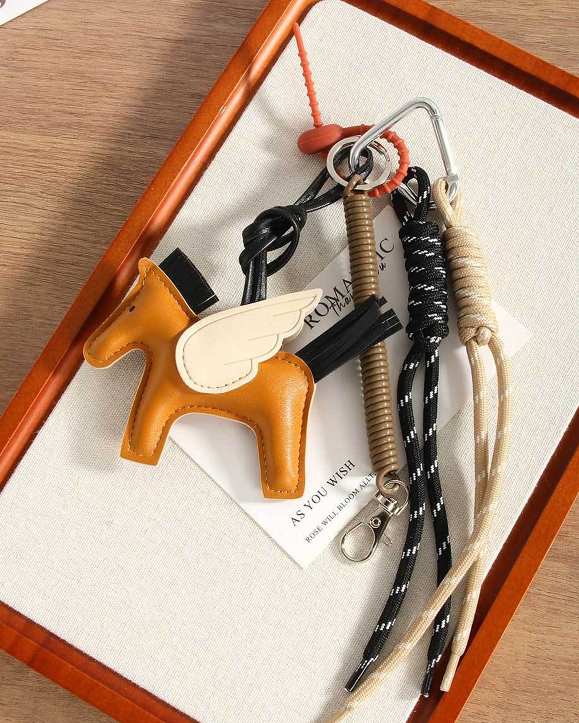 The Faire Pegasus Keychain features a brown PU leather winged horse charm, black and tan rope loops, a metal clip, and a whistle, resting on a tray with paper underneath.