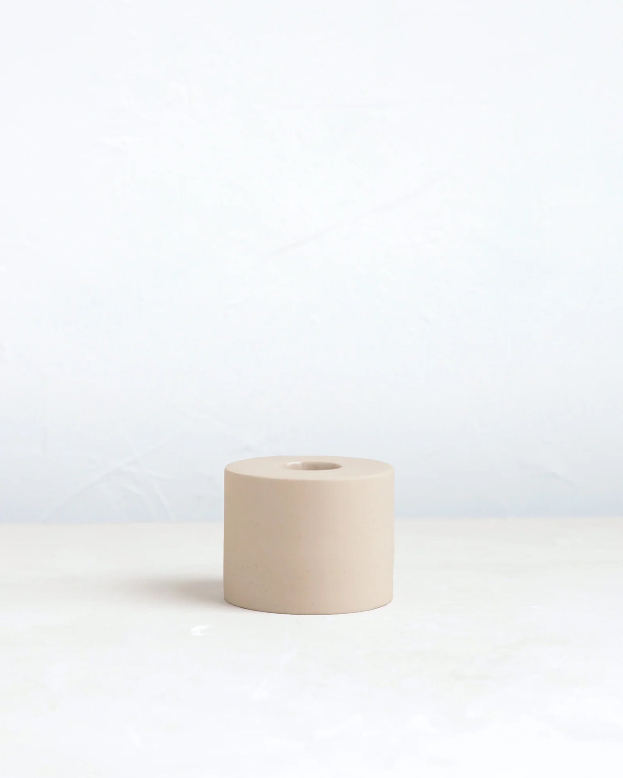 The Floral Society's Petite Taper Holder is a small, cylindrical beige ceramic piece with a center hole, ideal for displaying taper candles in modern spaces. Shown on a light surface with a plain white background.