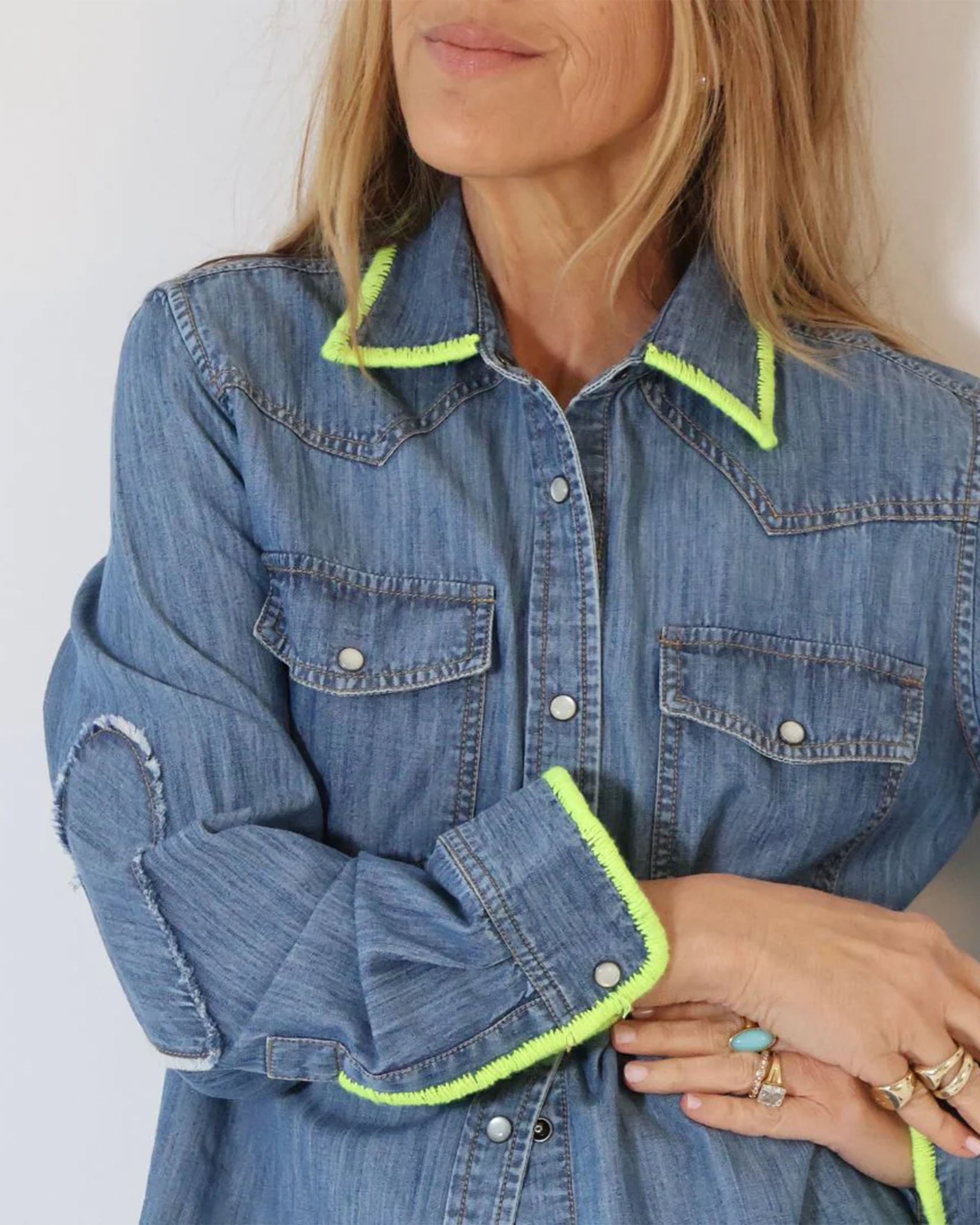 A woman stands with folded arms, her face partially visible, wearing the Kerri Rosenthal Pia Denim Western Shirt featuring bright yellow-green trim on the collar and cuffs, and light-colored rings on her fingers.