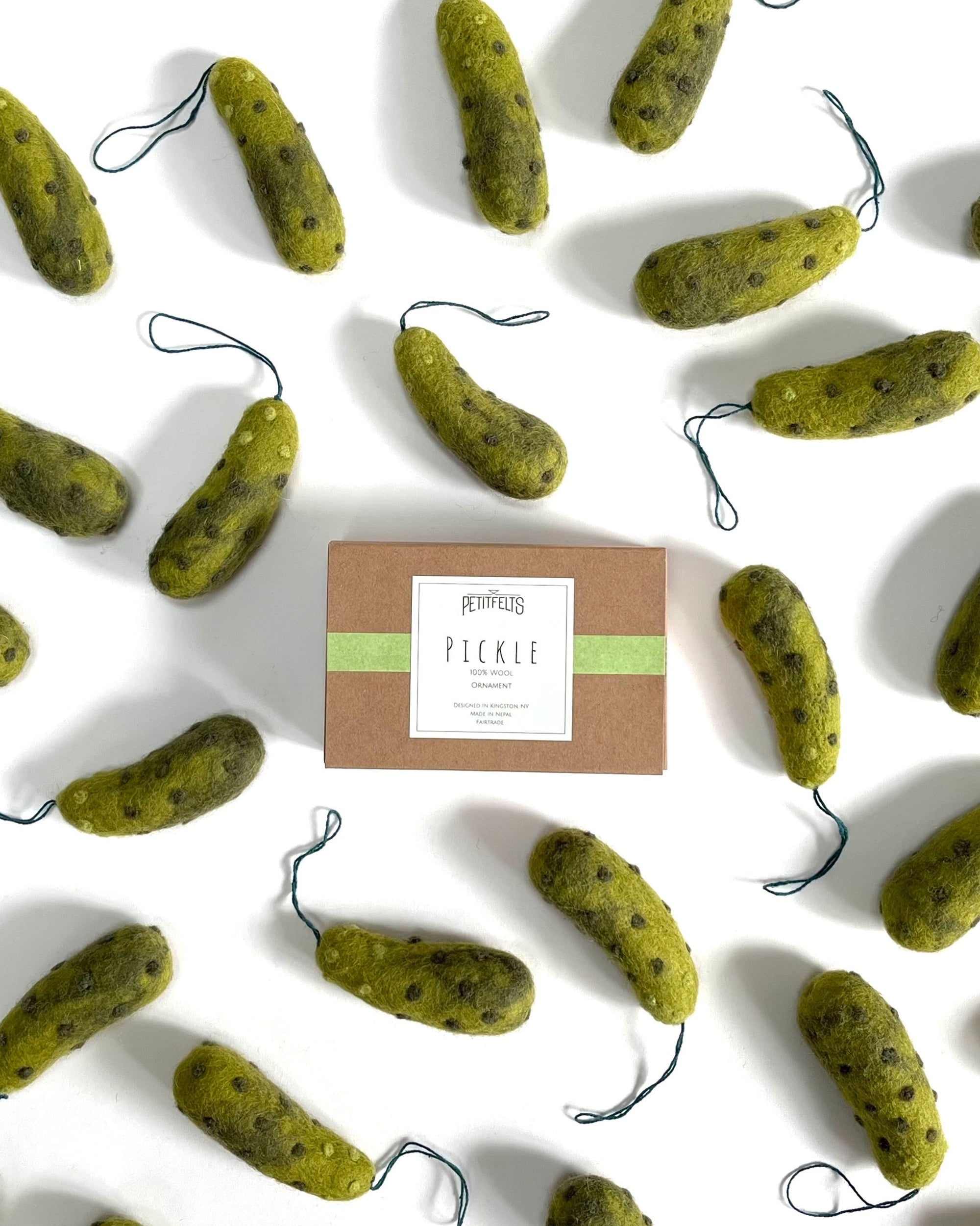 A box labeled "Pickle" is surrounded by several green Faire Felted Ornaments shaped like pickles, each with a loop for hanging, all displayed on a white background.