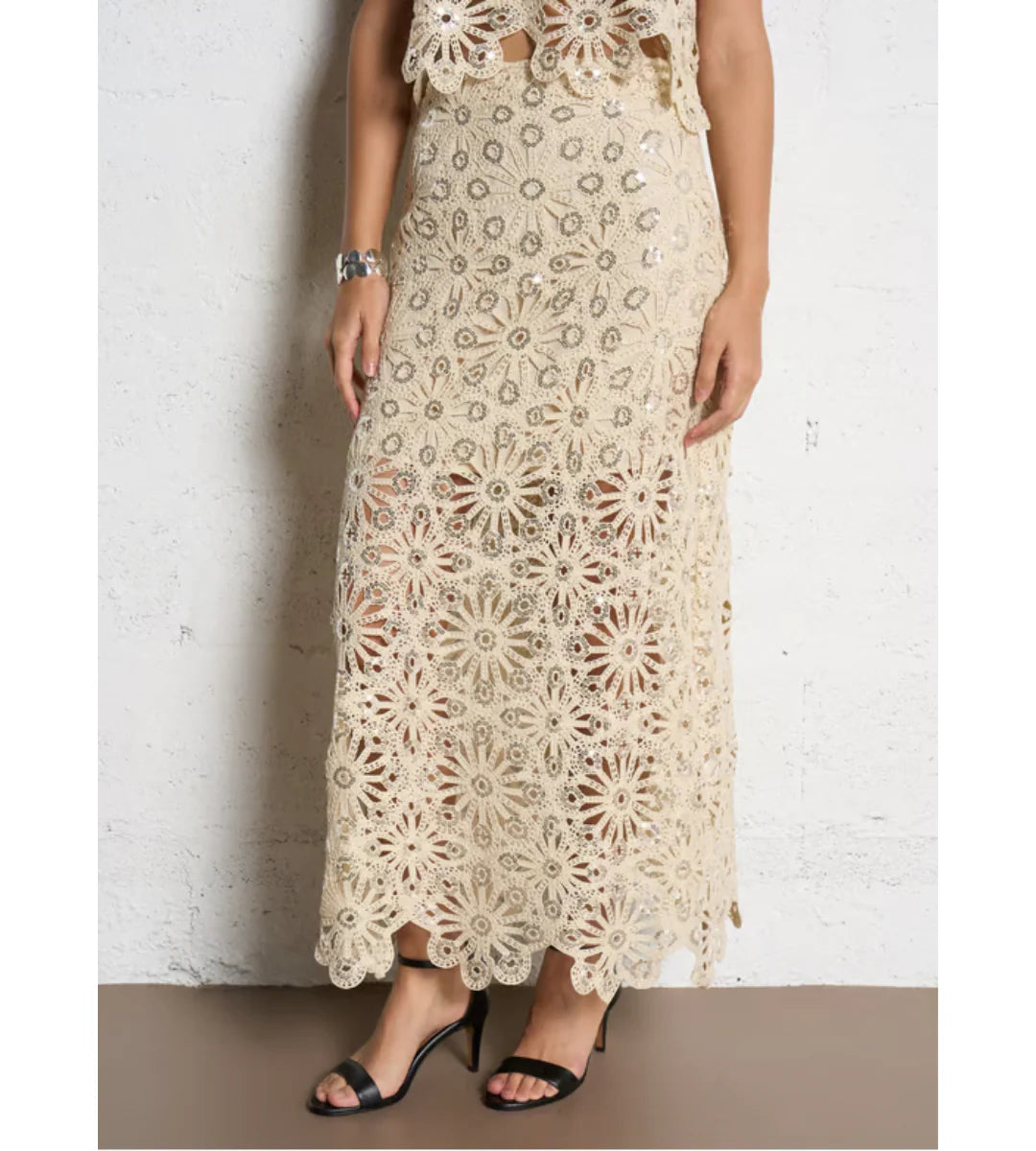 A person wearing the SUNCOO Fanny Skirt in beige crochet with floral patterns and lace detailing, paired with black heeled sandals and a silver bracelet, stands against a white textured wall.