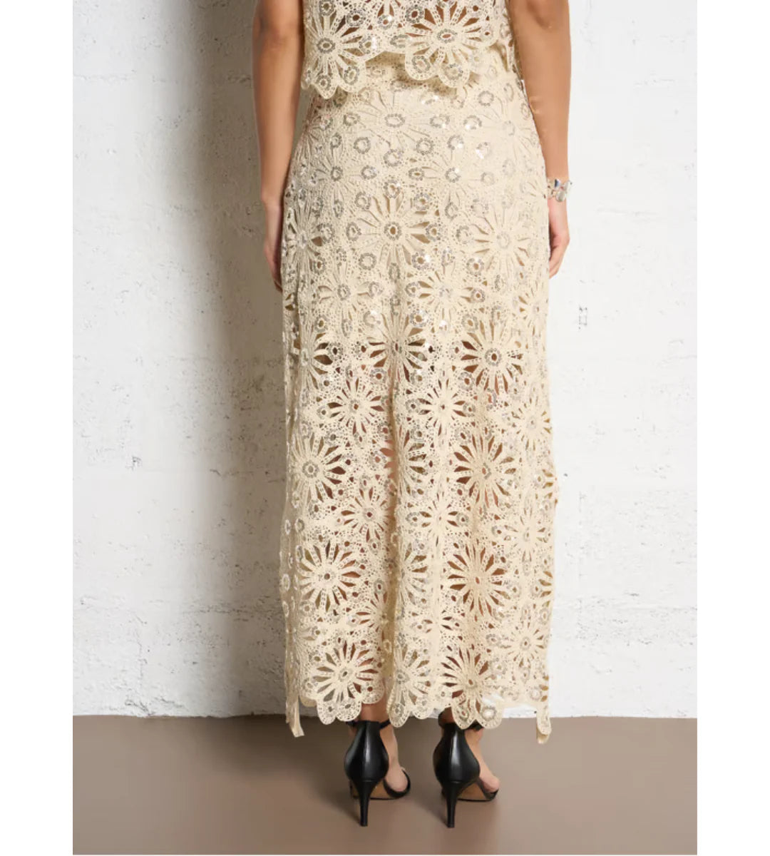 A person stands against a white textured wall wearing the SUNCOO Fanny Skirt in cream, featuring intricate lace and floral patterns. The semi-sheer skirt is elegantly paired with black high-heeled sandals.