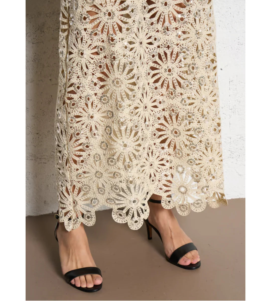 Close-up of a person wearing black open-toe heels and the SUNCOO Fanny Skirt, featuring intricate crochet floral lace, set against a white wall and brown floor.