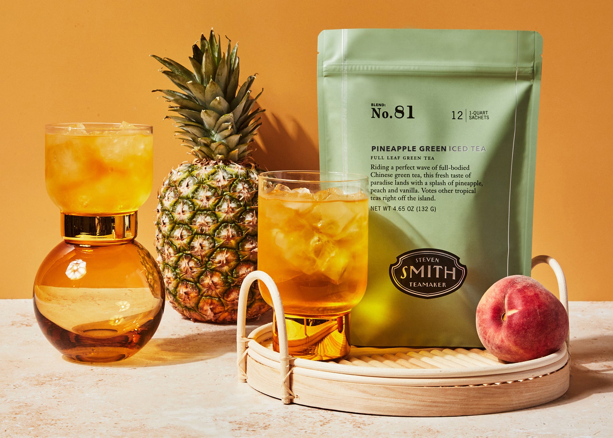 A glass and pitcher of iced tea sit on a tray with peach, pineapple, and mango, next to a green package labeled "Smith Tea" by Faire, all set against a warm yellow background.