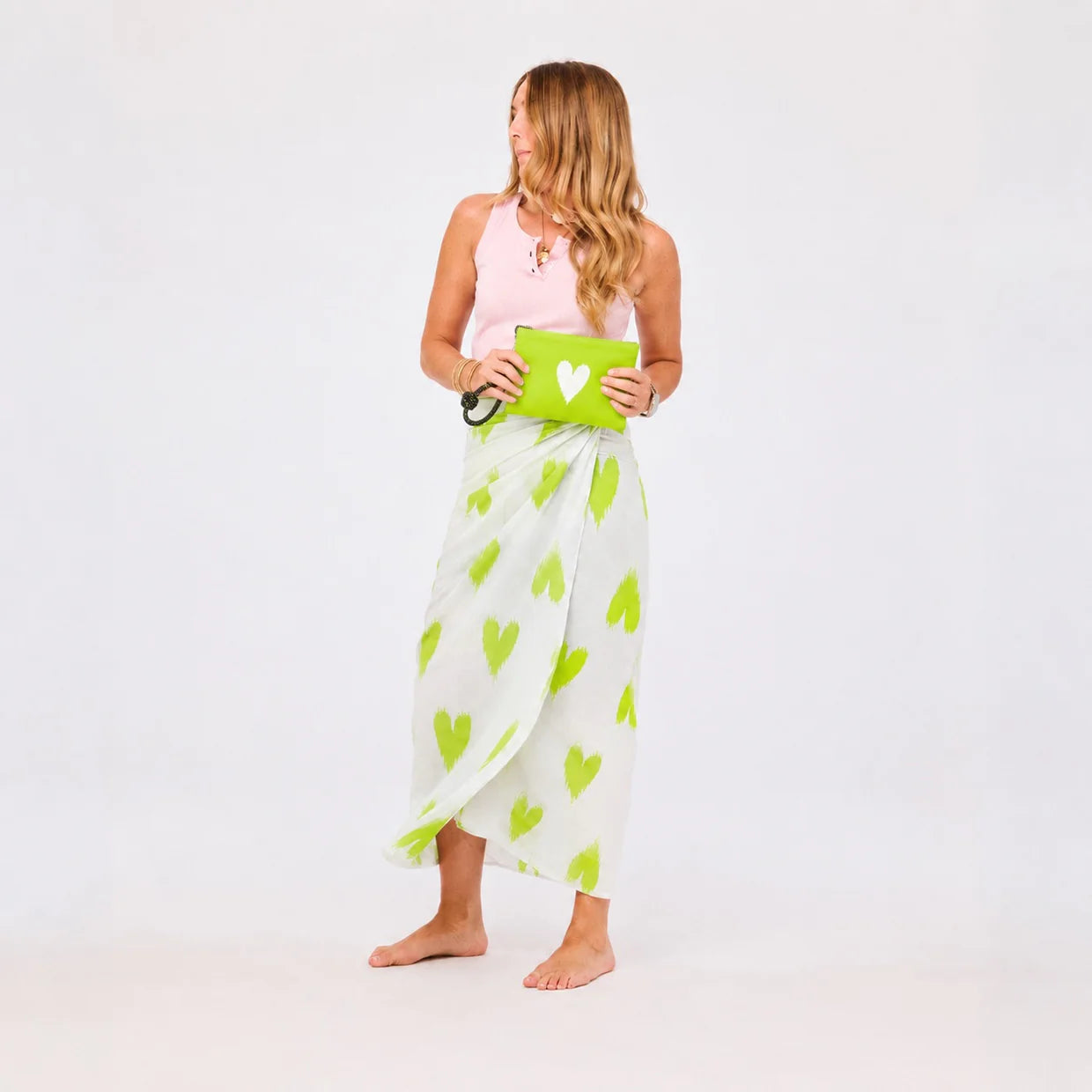 A woman with long, wavy hair wears a pink tank top and a white wrap skirt with green heart prints. She holds the Kerri Rosenthal KR Heart Wristlet and stands barefoot against a plain white background.
