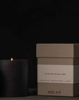 A black ADLAN Candle with a lit flame is positioned next to its open lid and a beige box marked "Playing with Fire" by ADLAN Fragrances. The packaging emits hints of smoky birch, Japanese ember, frankincense, and allspice, enhanced by the dark backdrop.