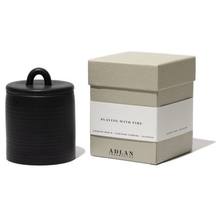 A black ADLAN candle with a lid is placed next to a beige box labeled &quot;ADLAN Candle&quot; by ADLAN Fragrances. The packaging details the scent profile, highlighting smoked birch, Turkish oud, agarwood, and a hint of jalapeño.