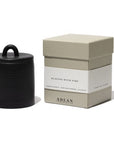 A black ADLAN candle with a lid is placed next to a beige box labeled "ADLAN Candle" by ADLAN Fragrances. The packaging details the scent profile, highlighting smoked birch, Turkish oud, agarwood, and a hint of jalapeño.