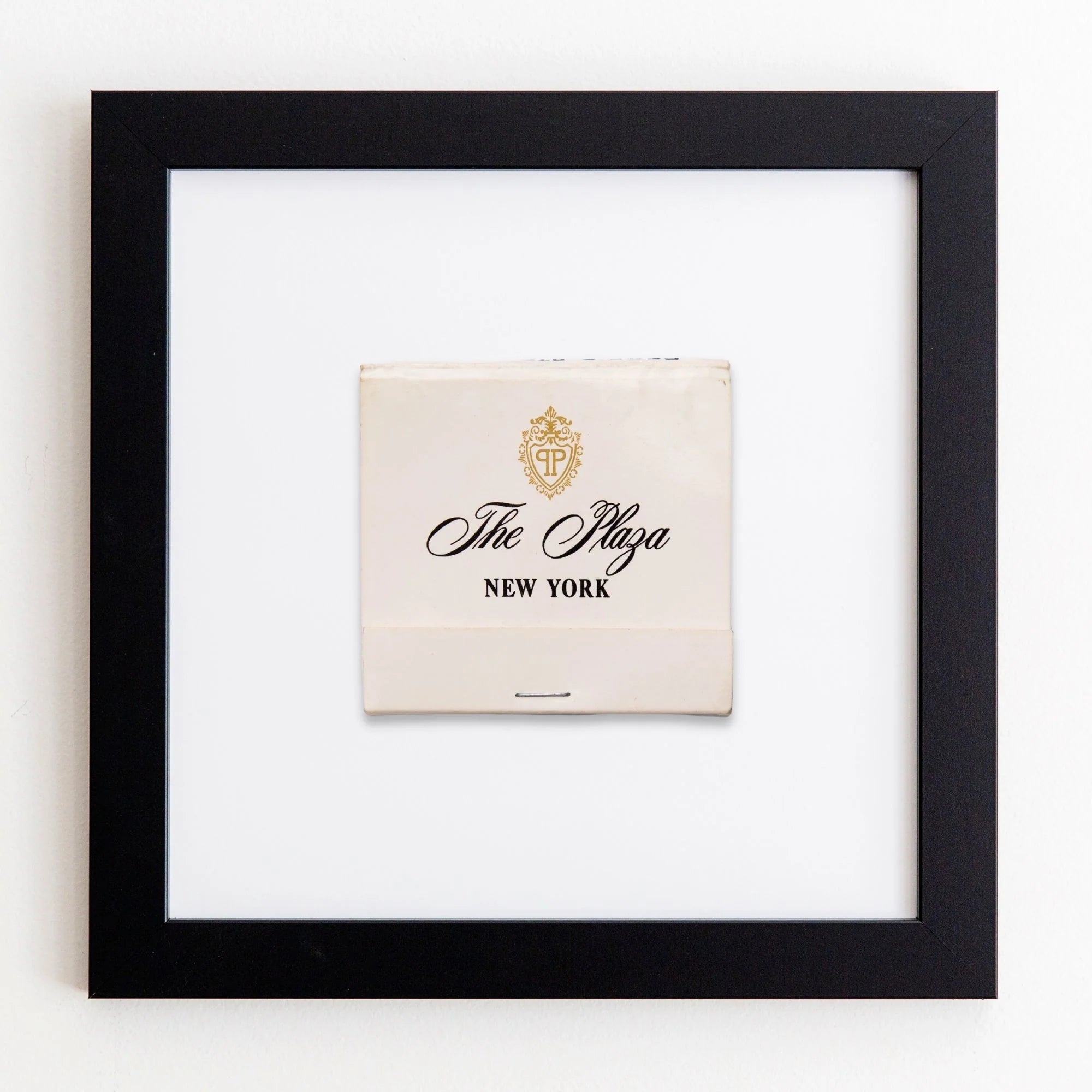 A framed matchbook from The Plaza Hotel, New York, displayed against a white wall. The matchbook, in off-white color, features the hotel’s logo and name in elegant typography within an Art Square Black Frame by Match South.